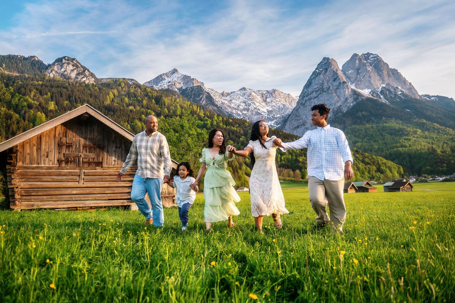 Happy family walking hand-in-hand on green meadow with Alpine mountains and wooden barn in background.