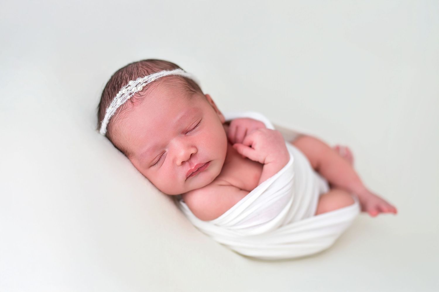 Sleeping newborn with sparkly headband wrapped in white fabric.