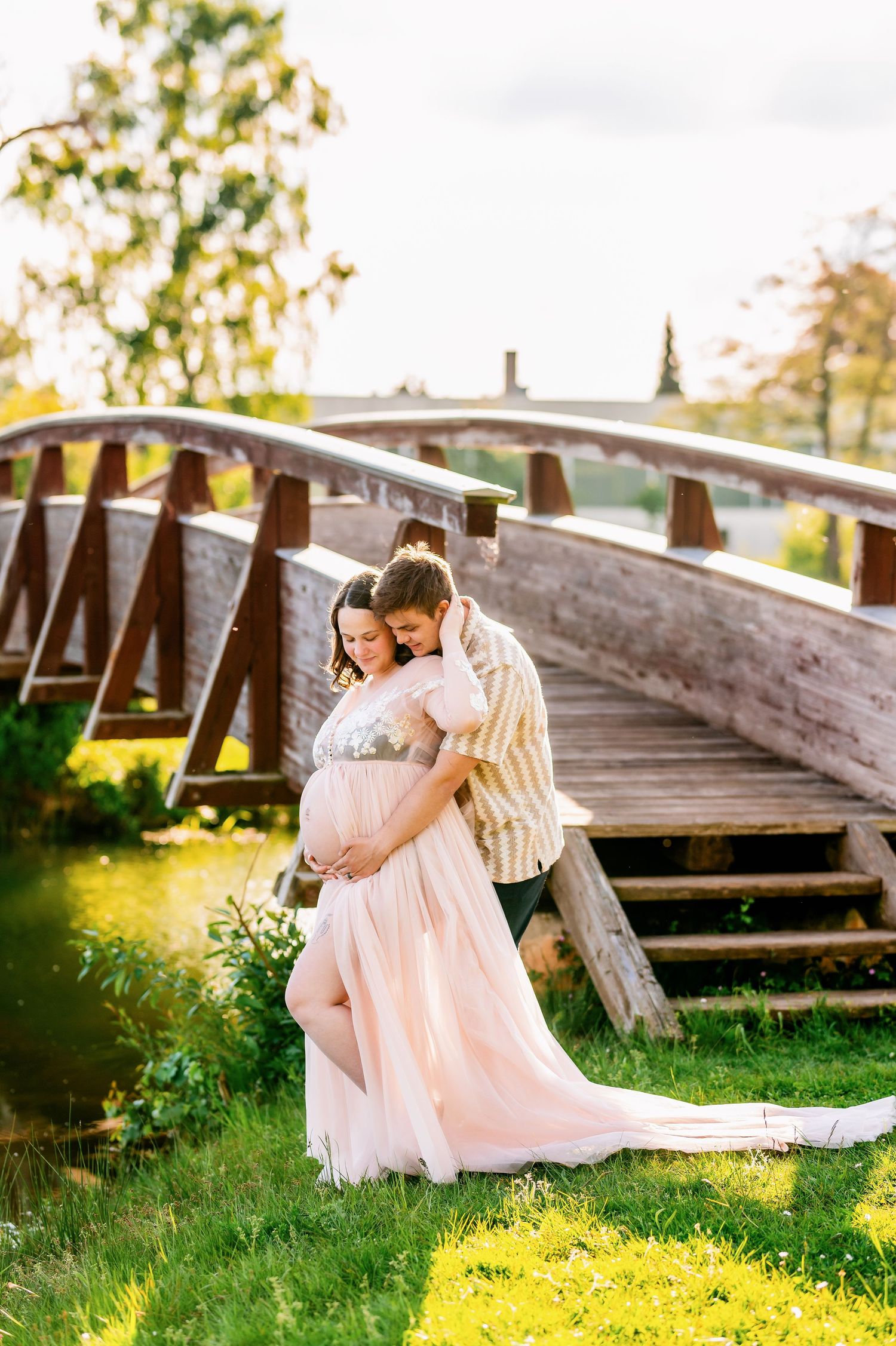 Romantic maternity photo of couple embracing on wooden bridge during golden hour.