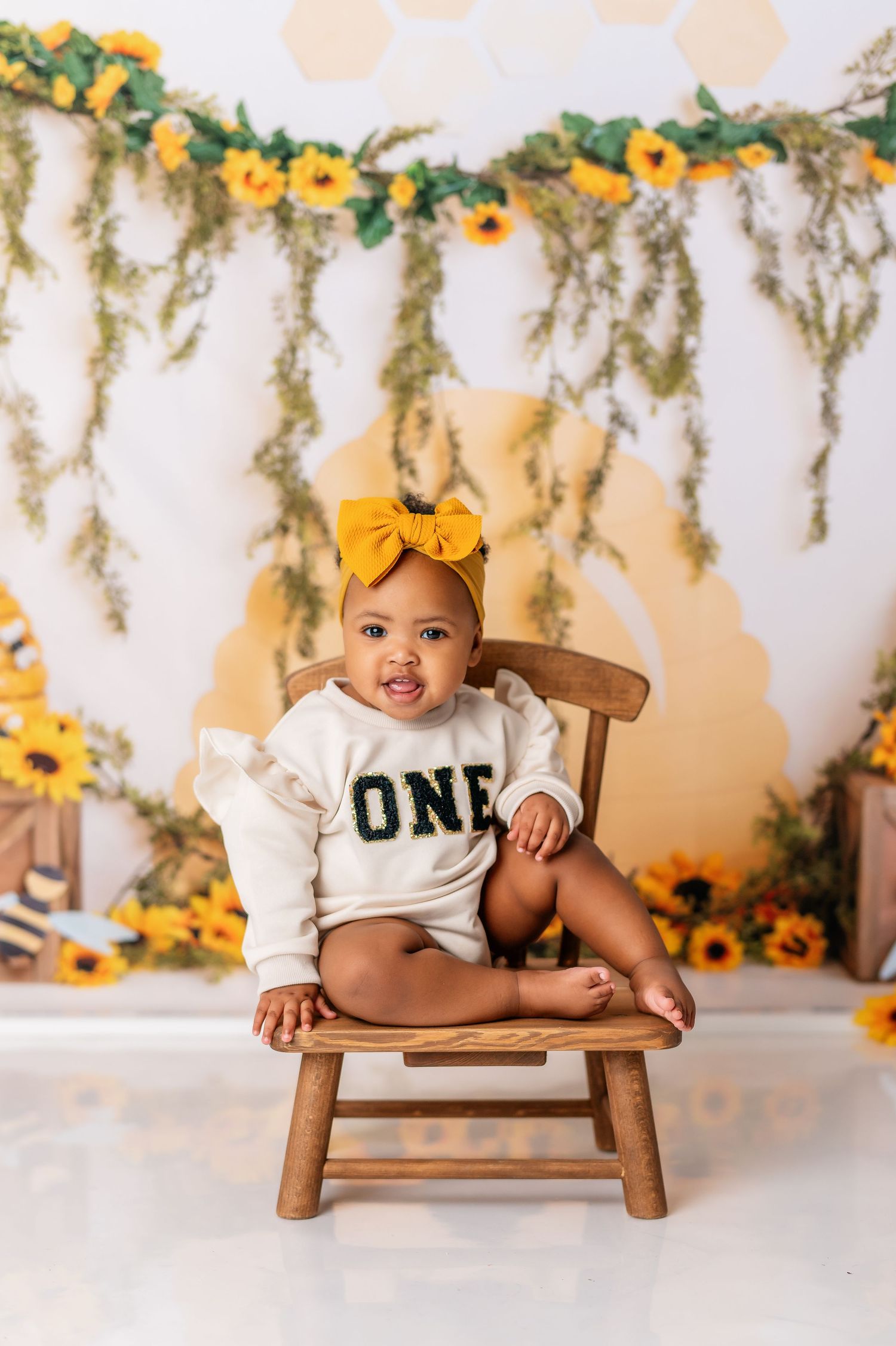 First birthday photo featuring wooden bench and sunflower themed backdrop with green garlands.