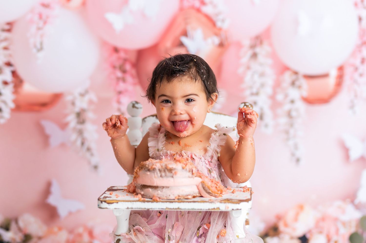 Pink and white balloon decorations create a festive backdrop for a cake smash photoshoot with pink frosting and sprinkles.