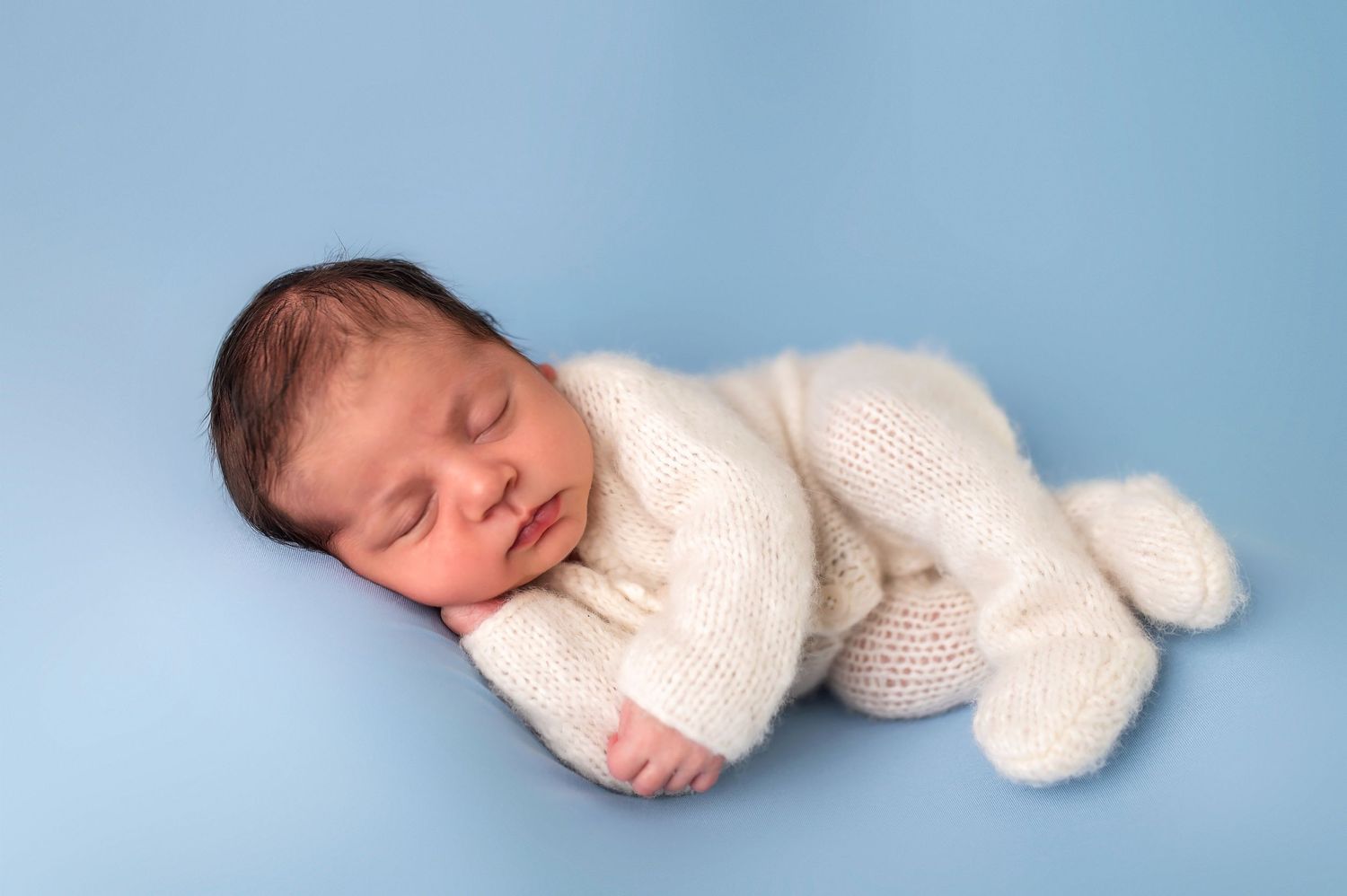 Sleeping newborn wrapped in white knit blanket on blue background.