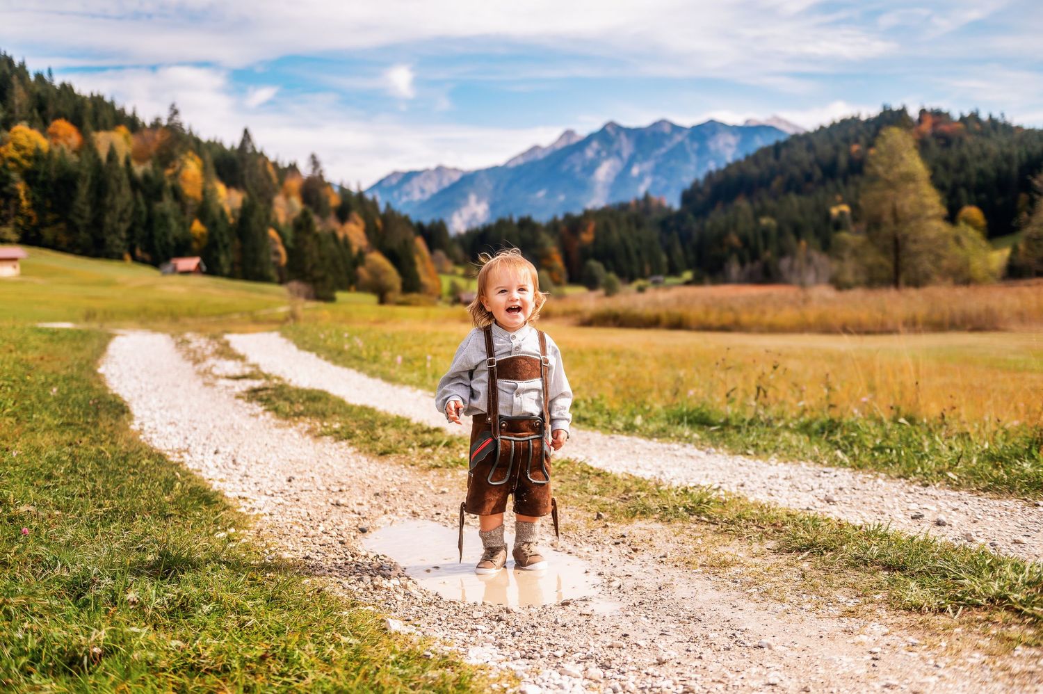 A cheerful youngster in traditional Bavarian attire stands on a gravel path with majestic Alps and fall colors in background.