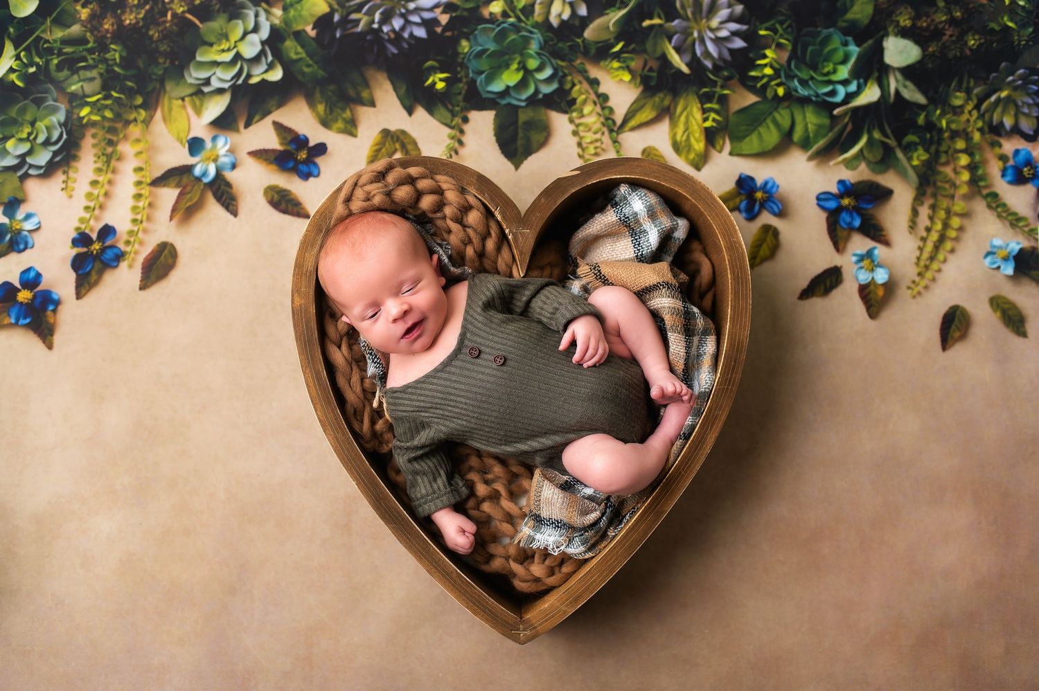 Sleeping newborn in wooden heart container surrounded by blue floral decorations.