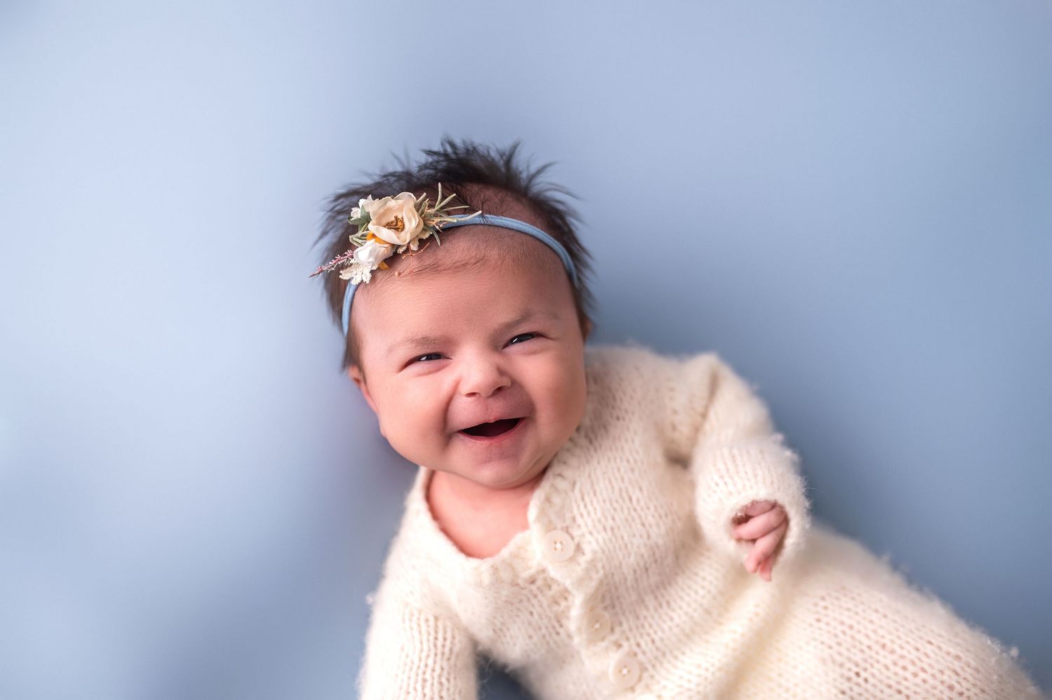 Baby in cream sweater with floral headband smiling against blue background.