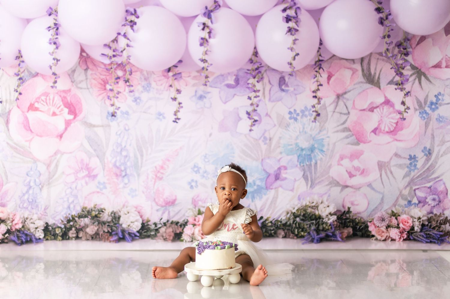 Pink and purple themed cake smash setup with balloons and floral backdrop for baby photography.