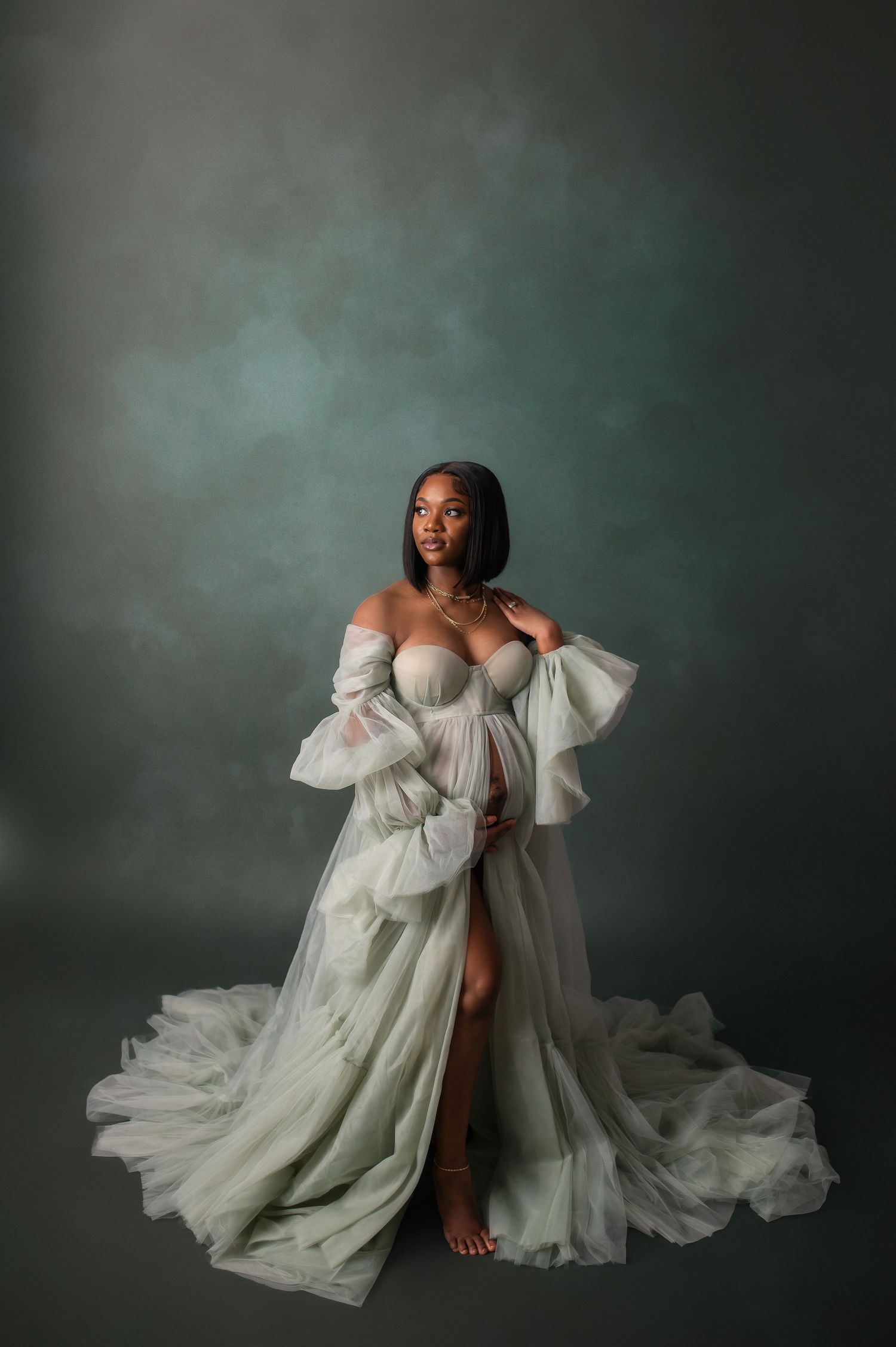 Ethereal maternity portrait in flowing white gown with dramatic side slit against moody backdrop.