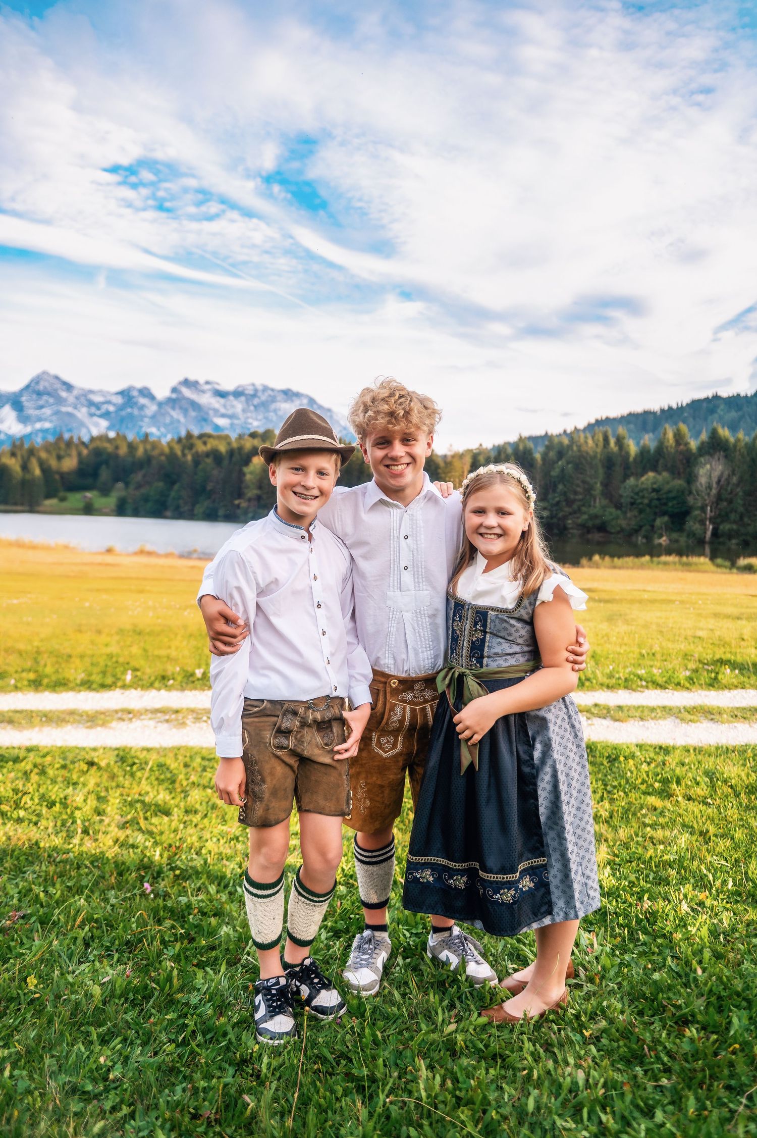 Three children in traditional Bavarian outfits play in a sunny mountain meadow with majestic Alps in the background.