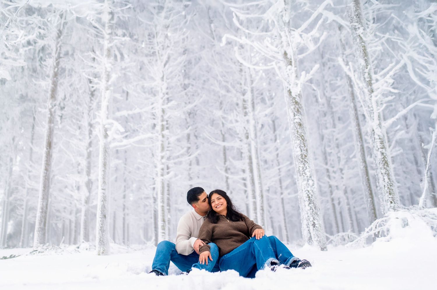 A couple sits together in a snowy forest with frost-covered trees creating a magical winter wonderland backdrop.