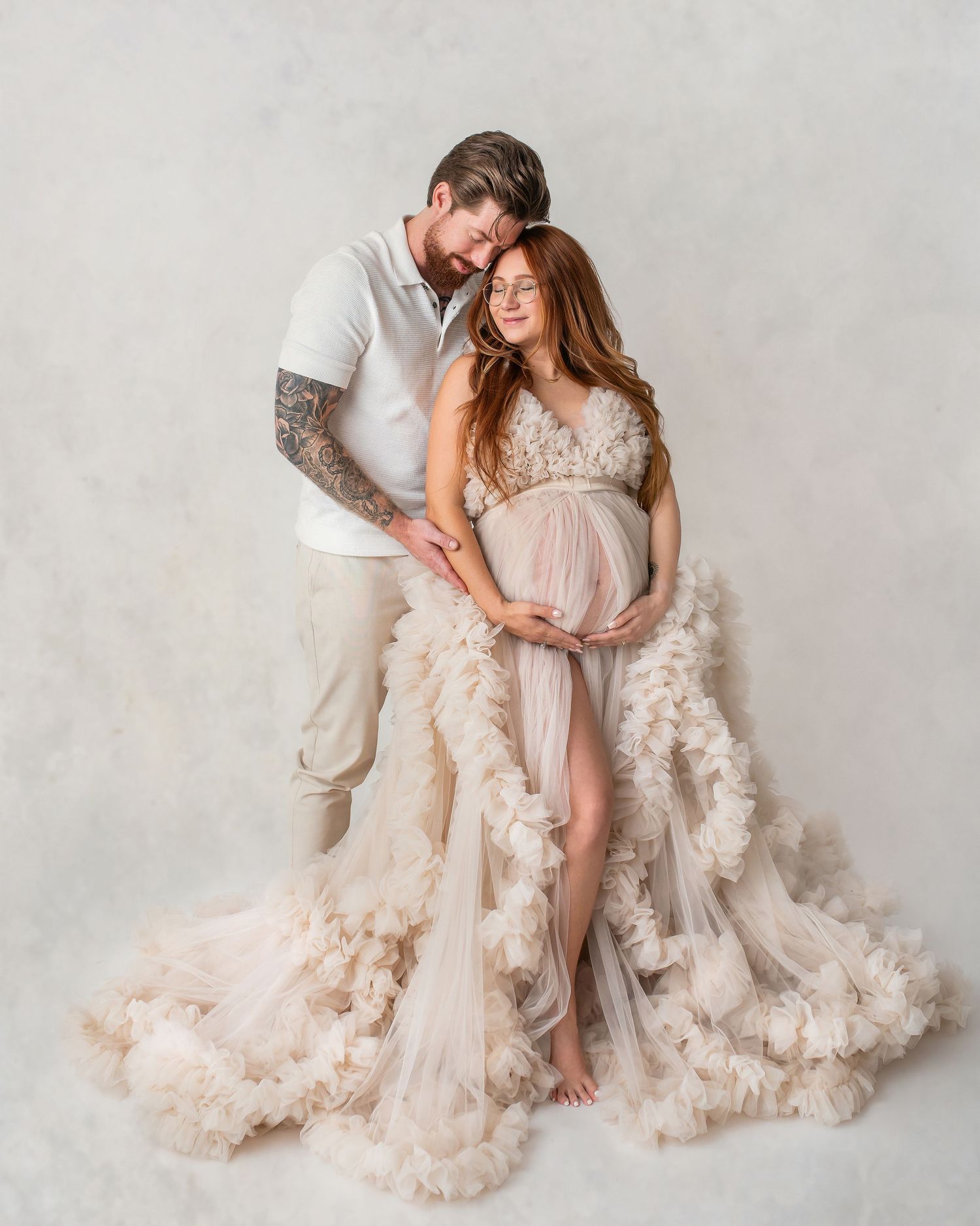 Couple in maternity photoshoot, woman in ruffled tulle gown cradling baby bump, man in white shirt embracing her tenderly.