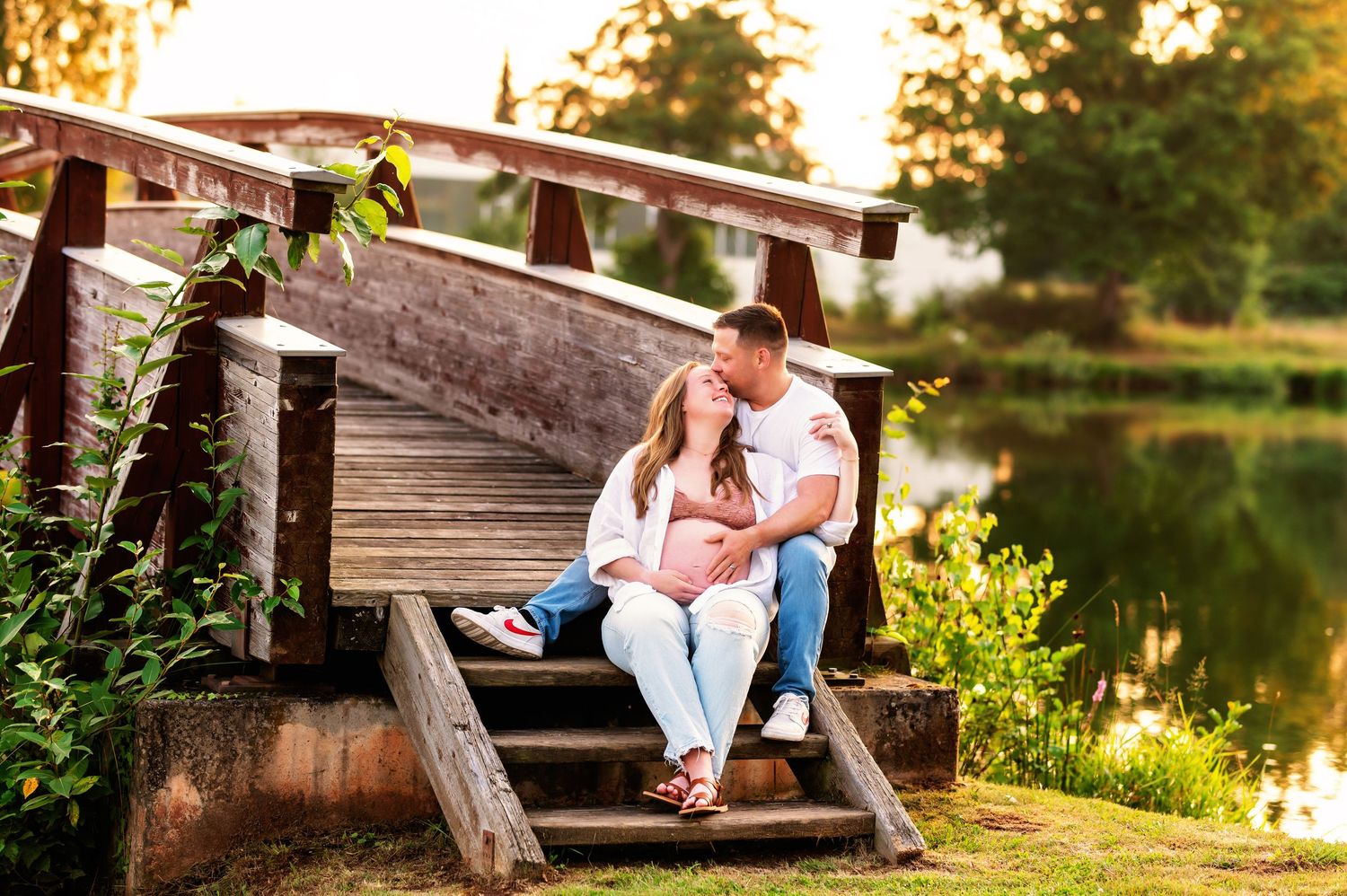 Couple sitting on wooden bridge steps by a pond, sharing a tender moment in a lush green outdoor setting.