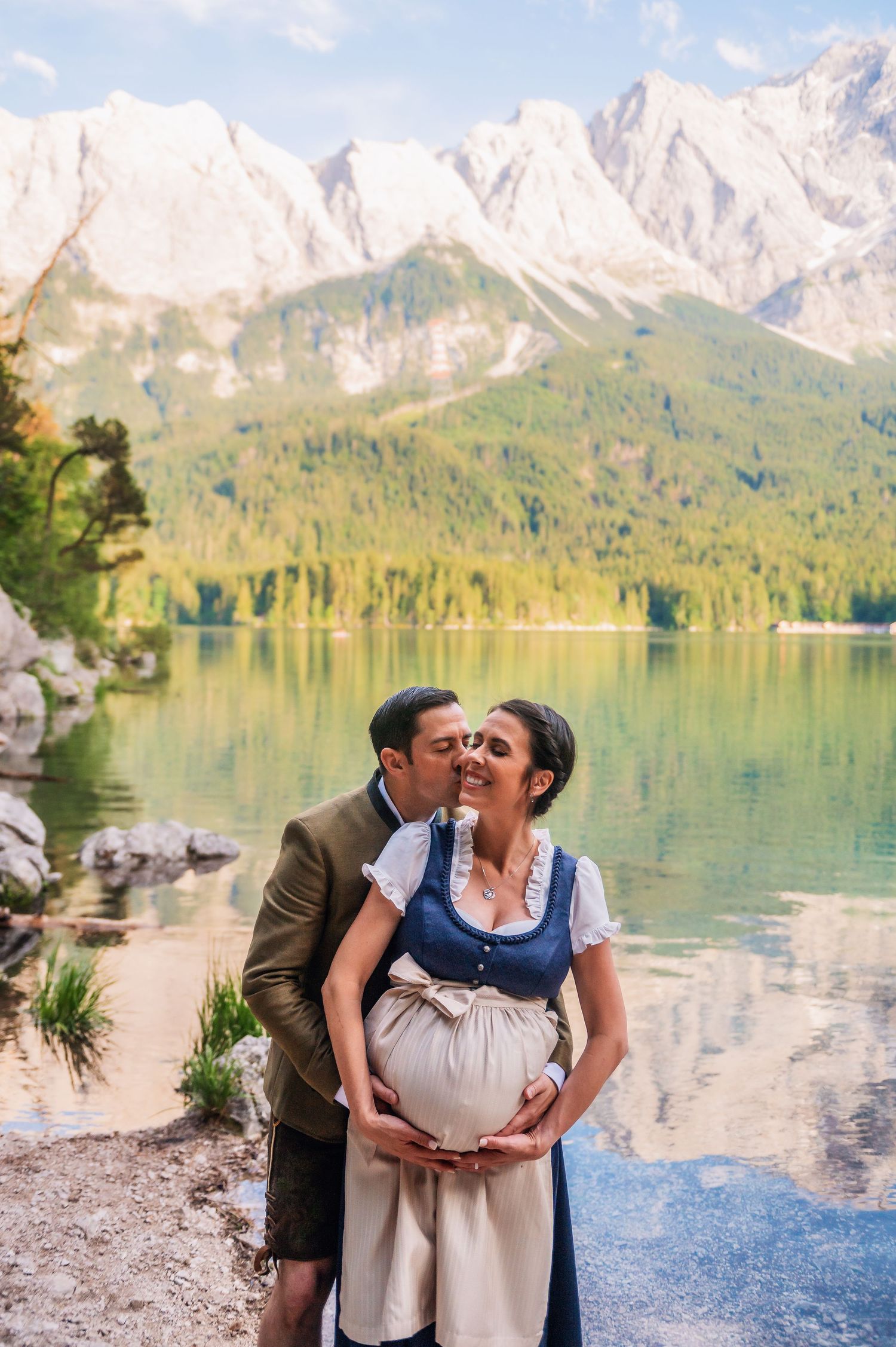 A romantic moment captured by an alpine lake with snow-capped mountains and evergreen forests in the background.