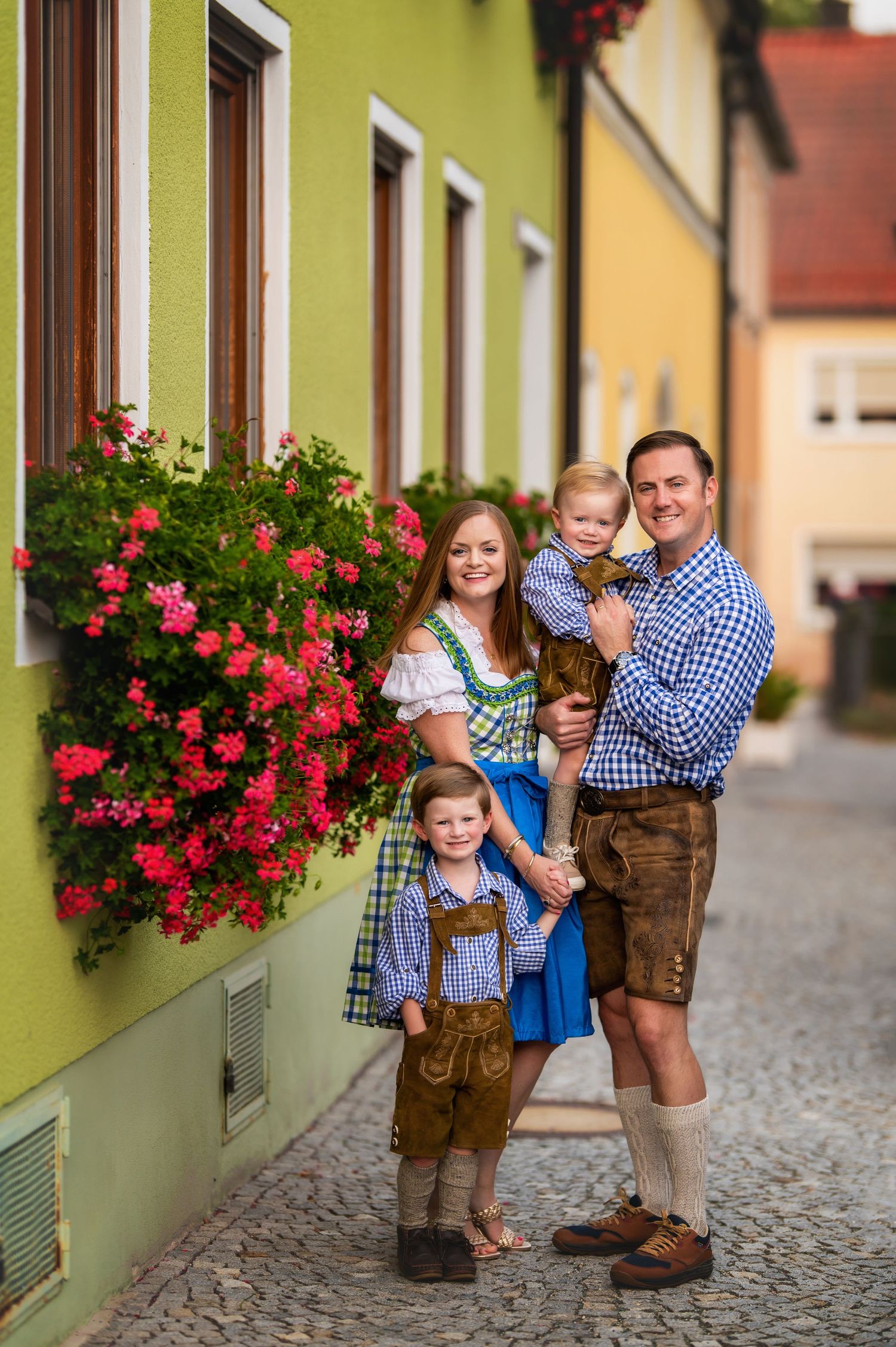 Traditional Bavarian family in dirndl and lederhosen poses by colorful building with pink flowers.