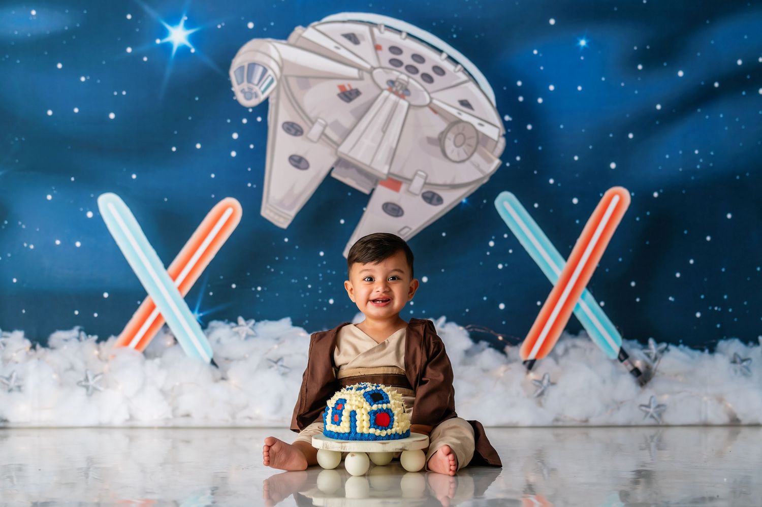 A baby dressed in Star Wars themed clothing sits with lightsabers and Millennium Falcon toys against a starry backdrop.