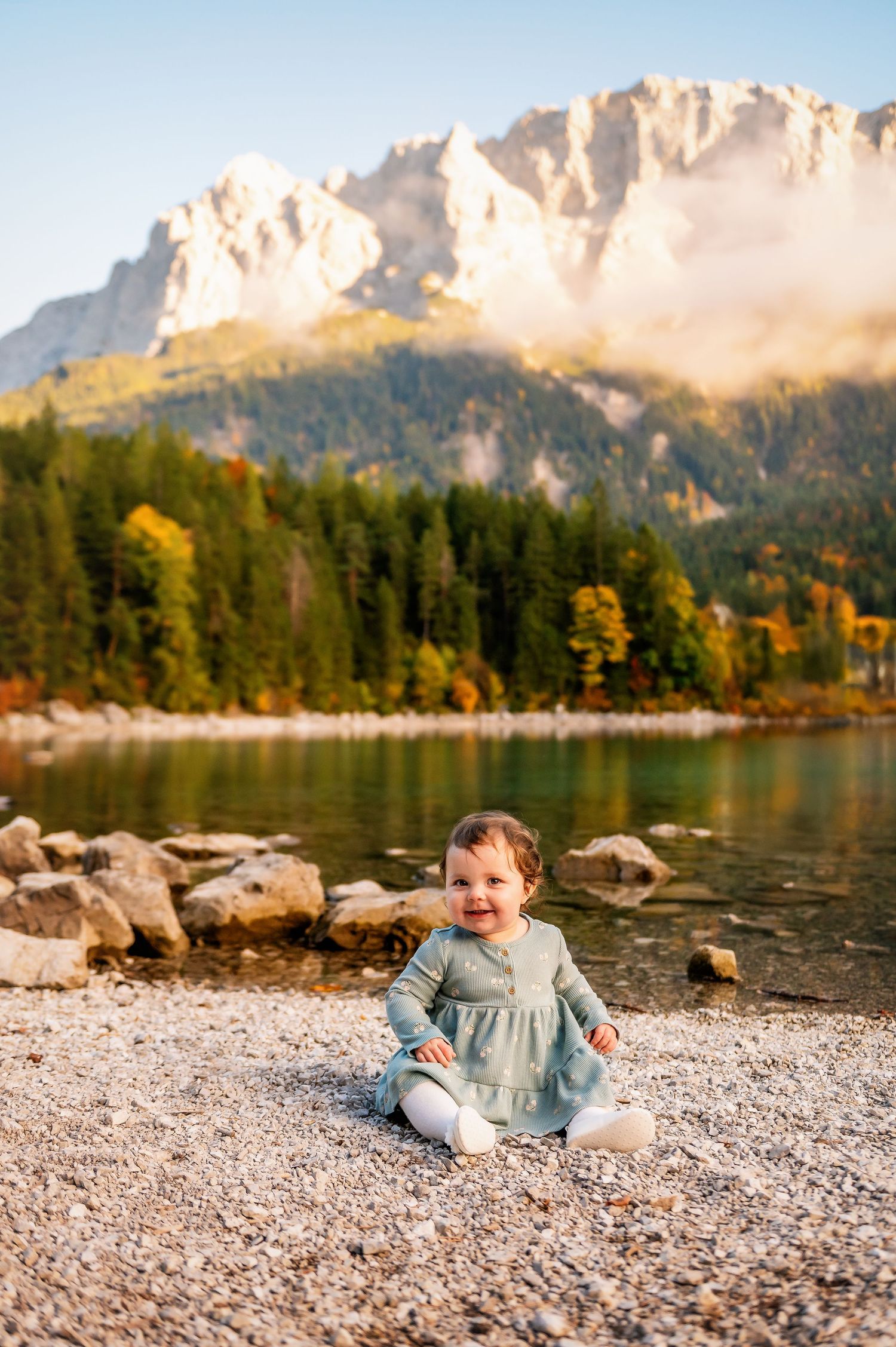 A scenic mountain lake shore with rocky beach and evergreen forest against rugged peaks during autumn.