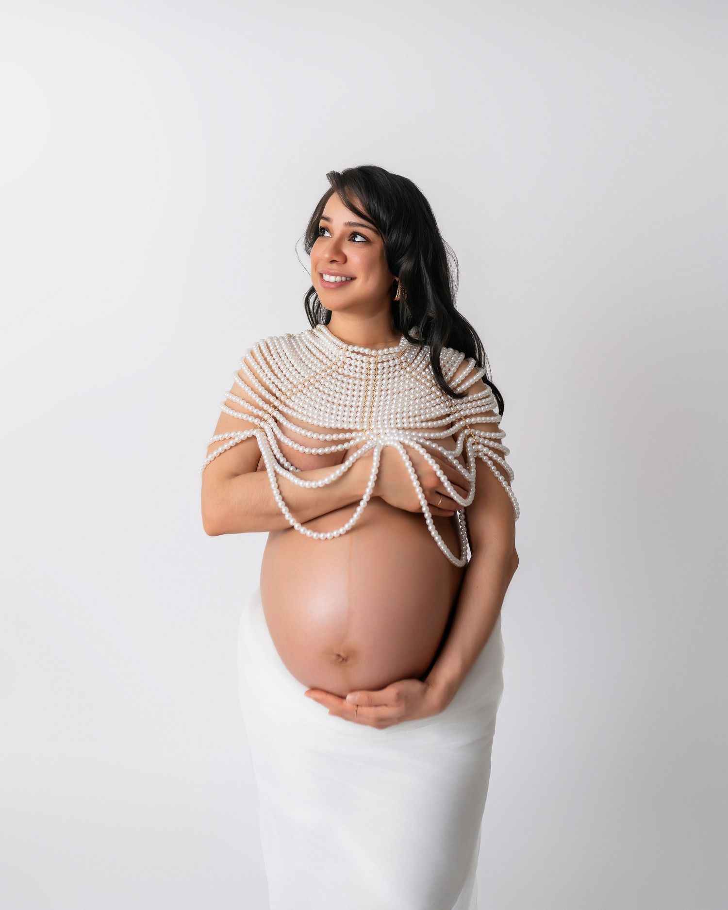 Pregnant woman wearing pearl body jewelry and white dress against white backdrop.