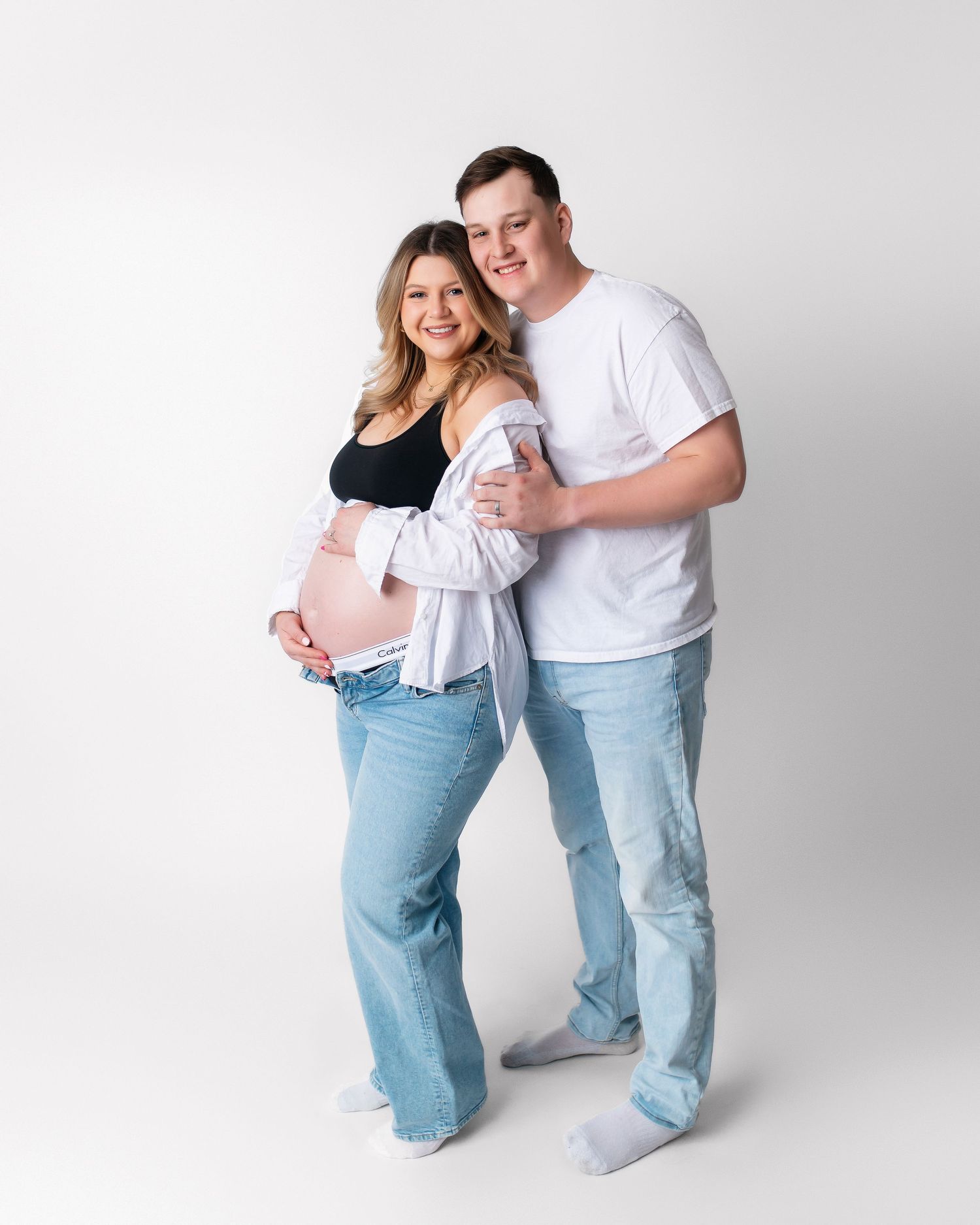 Expectant couple in casual attire embracing against white background.