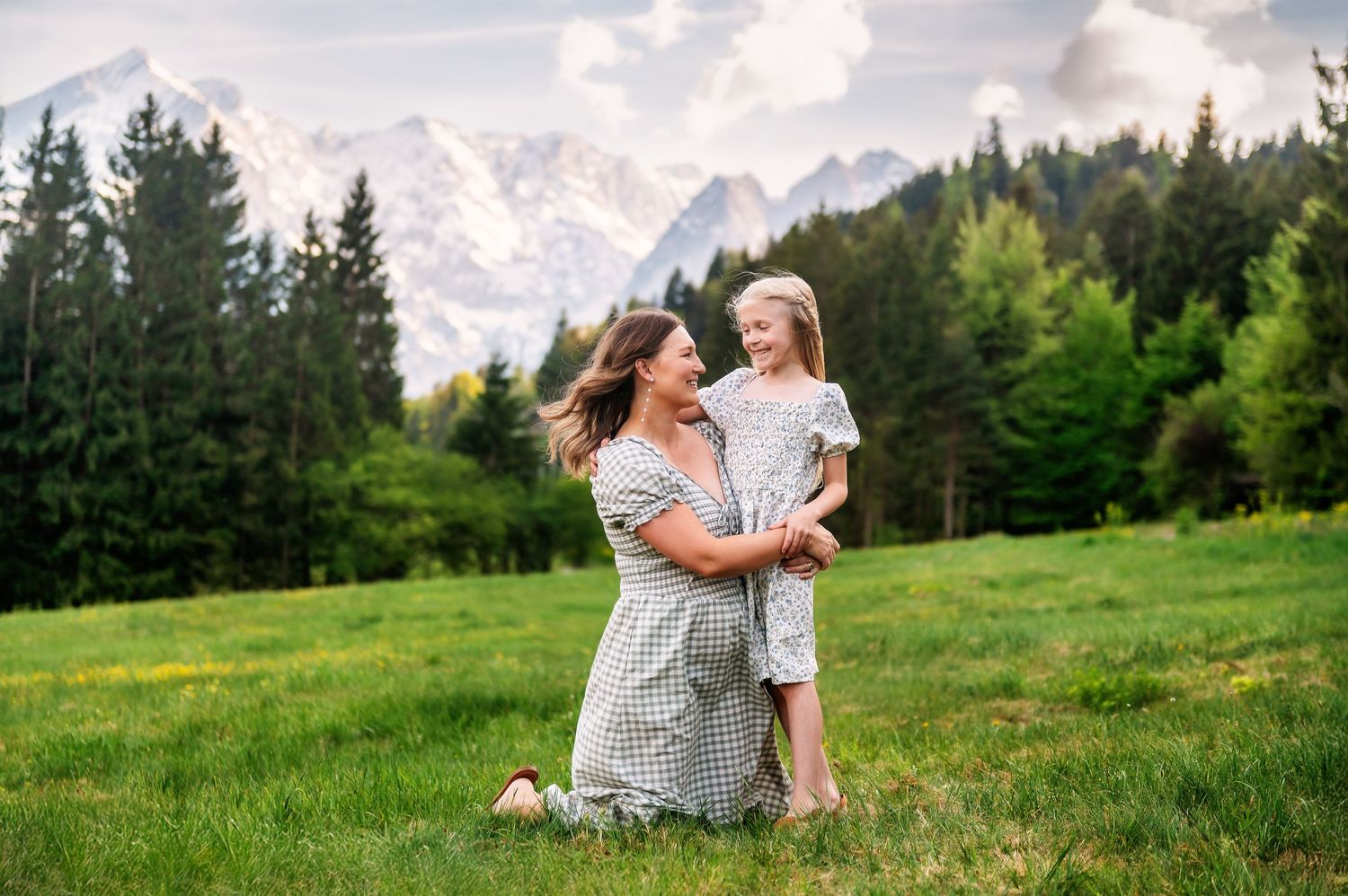 Mother and daughter embracing on green meadow with snow-capped mountains and pine forest backdrop.