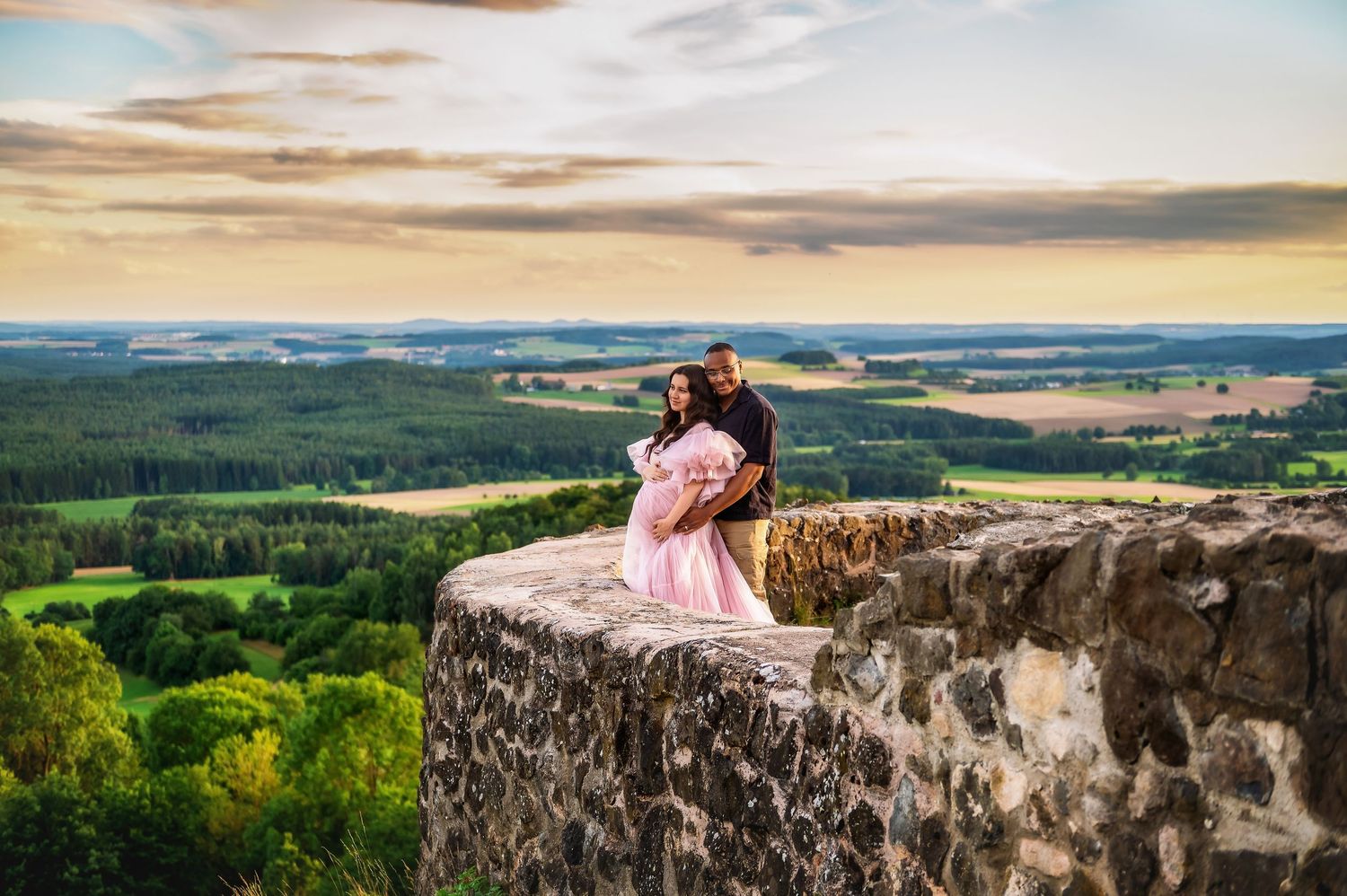 Couple posing on ancient stone castle wall overlooking lush green countryside landscape at sunset.