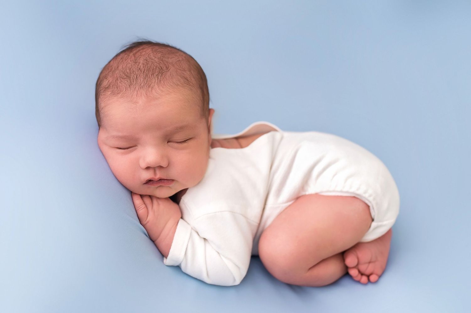 Sleeping baby in white onesie posed on soft blue background.