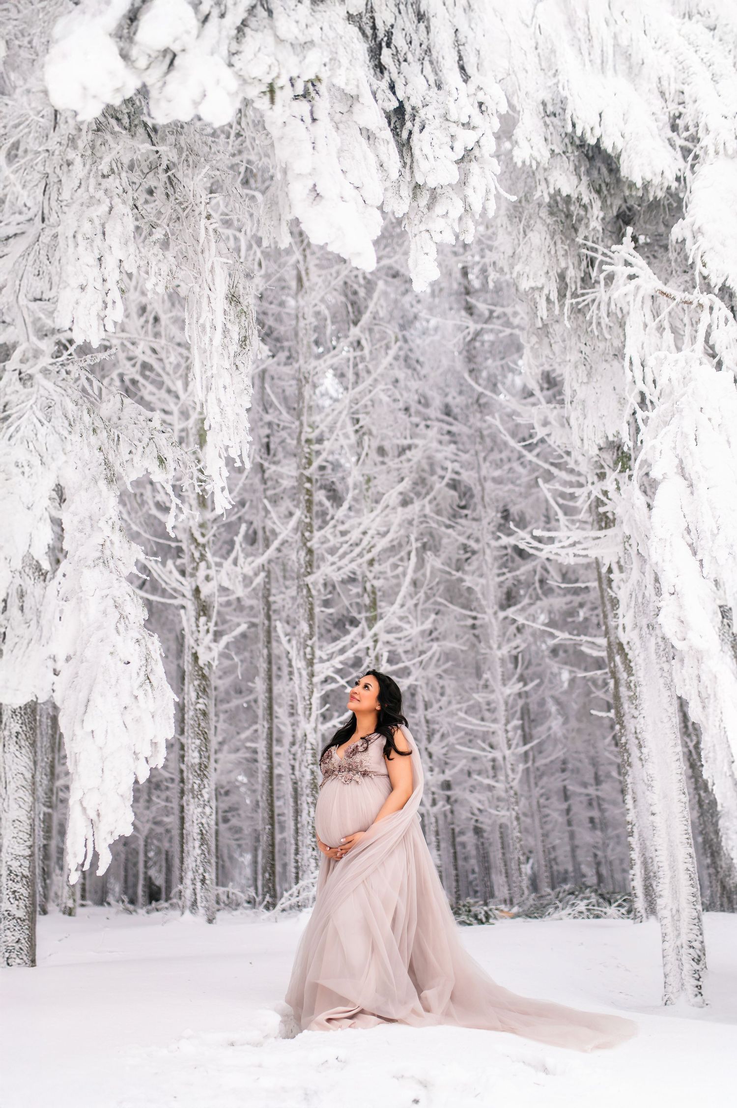 A flowing beige gown billows gracefully against a snowy winter forest backdrop with frost-covered trees.
