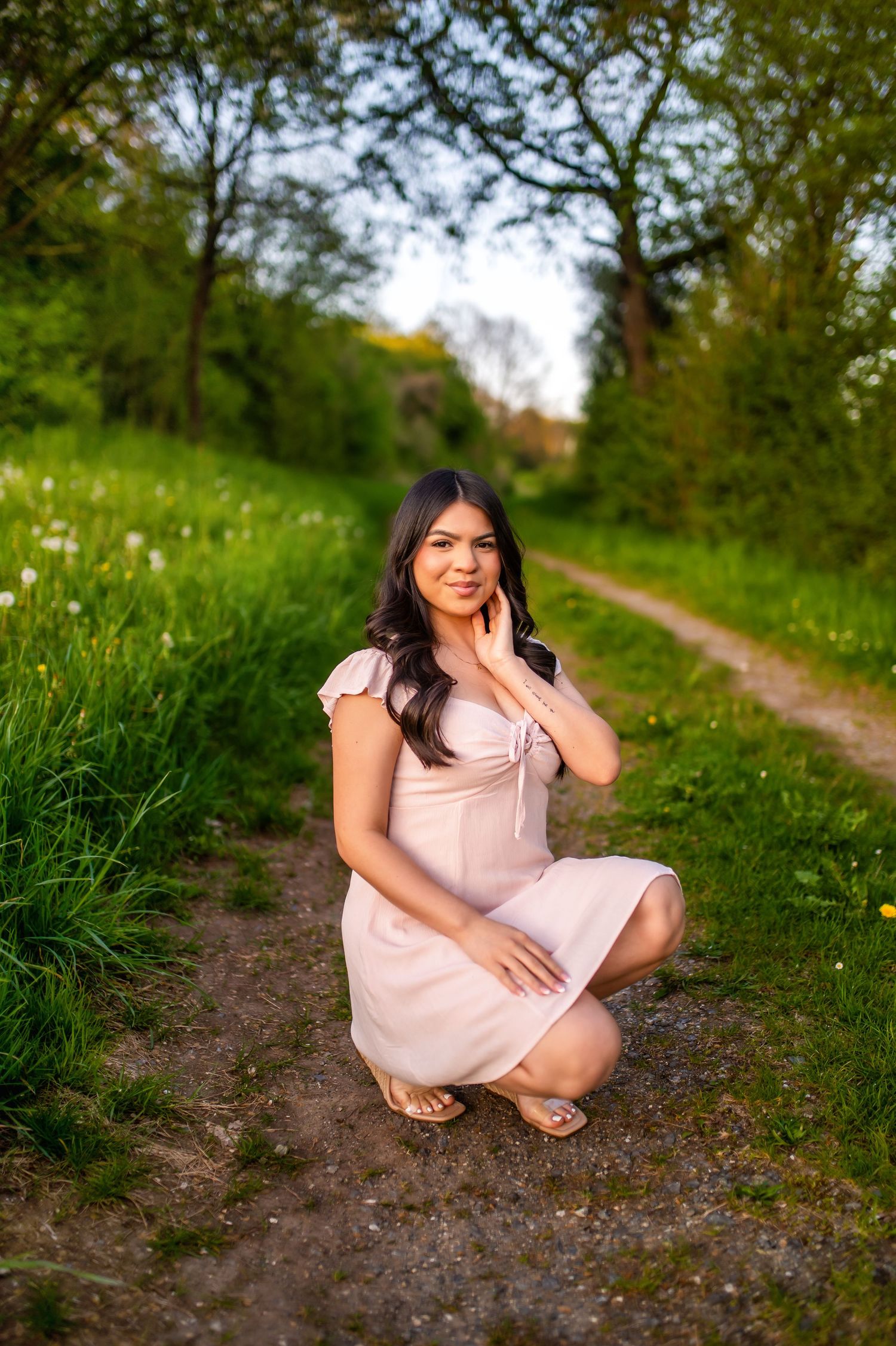Graduate in pink dress poses on rustic dirt path surrounded by spring greenery.