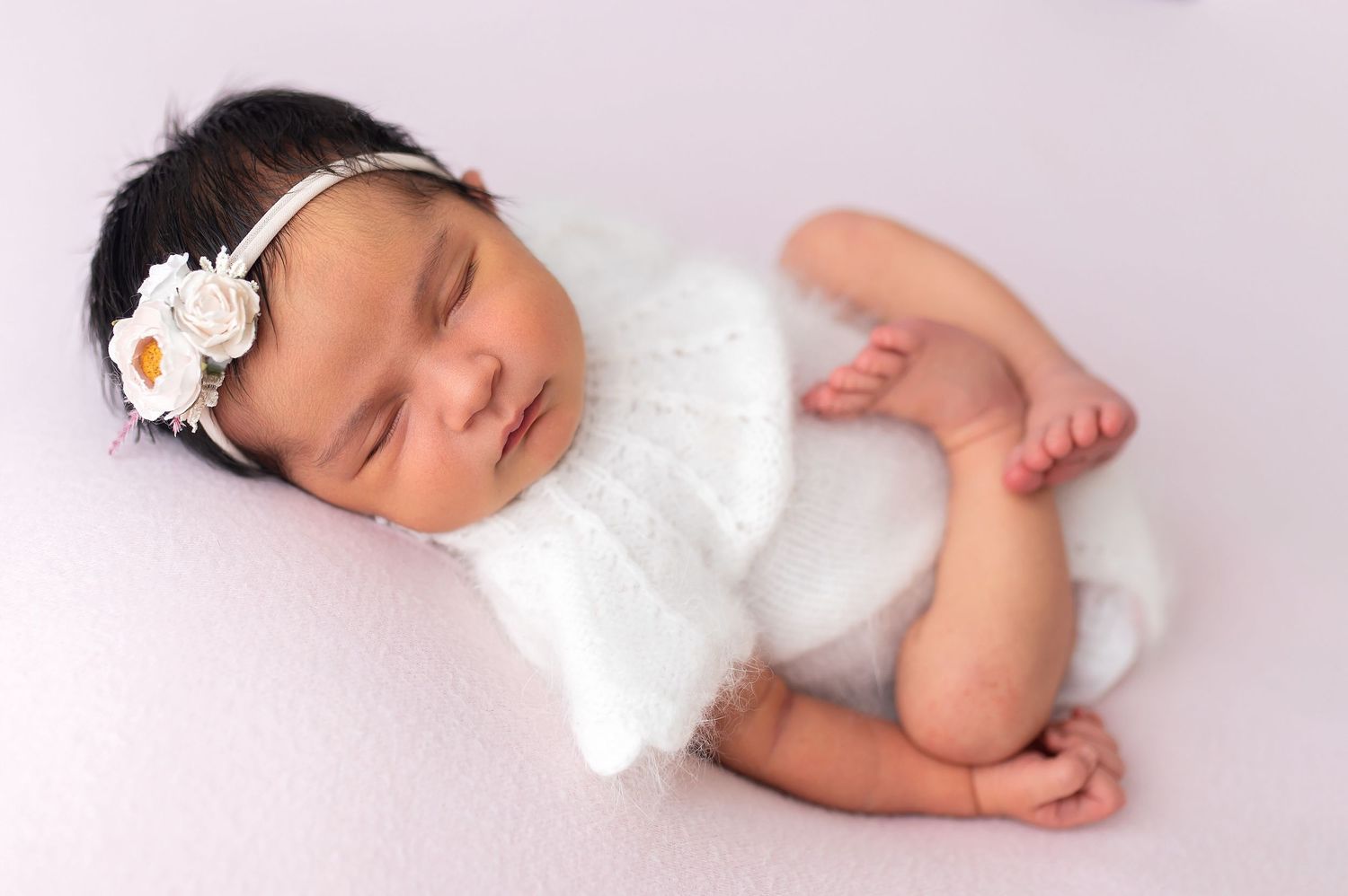Sleeping baby in white knit outfit with floral headband rests on pink background.