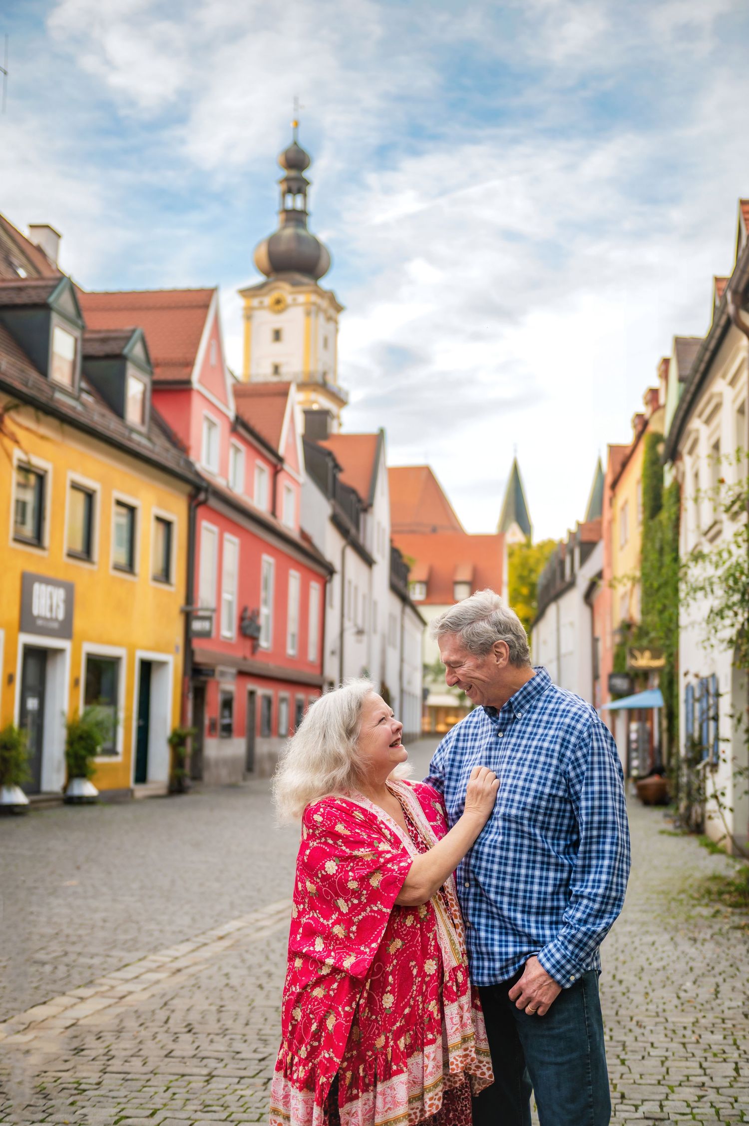 Romantic couple stands in picturesque European street with colorful buildings and church tower.