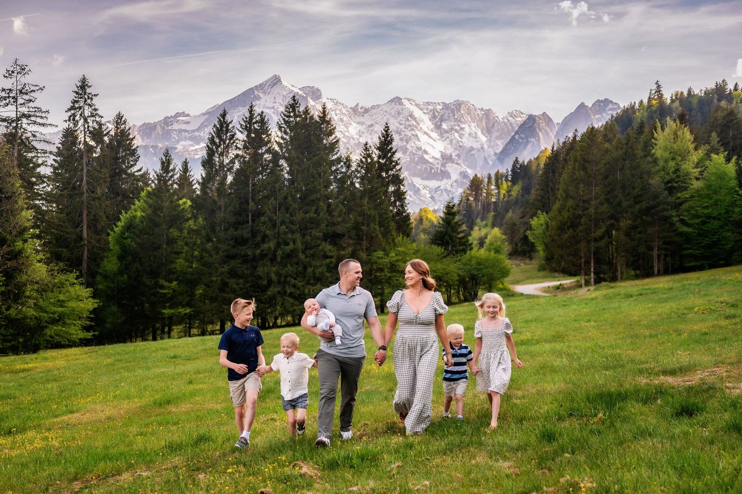 Family of five walking on green alpine meadow with snow-capped mountains and pine trees in background on sunny day.