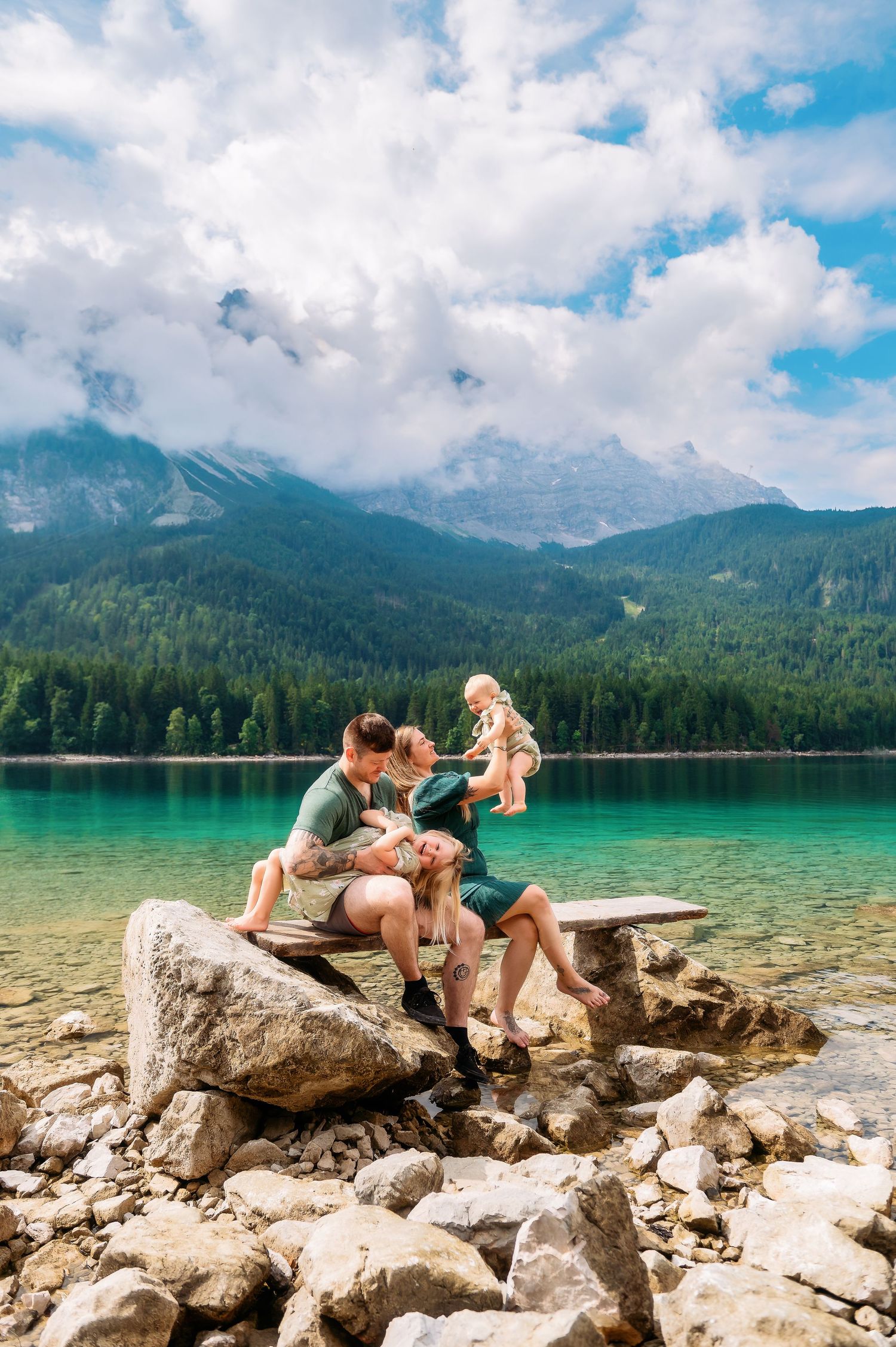 Family sitting on rocks by a turquoise alpine lake with mountains and forest in the background.