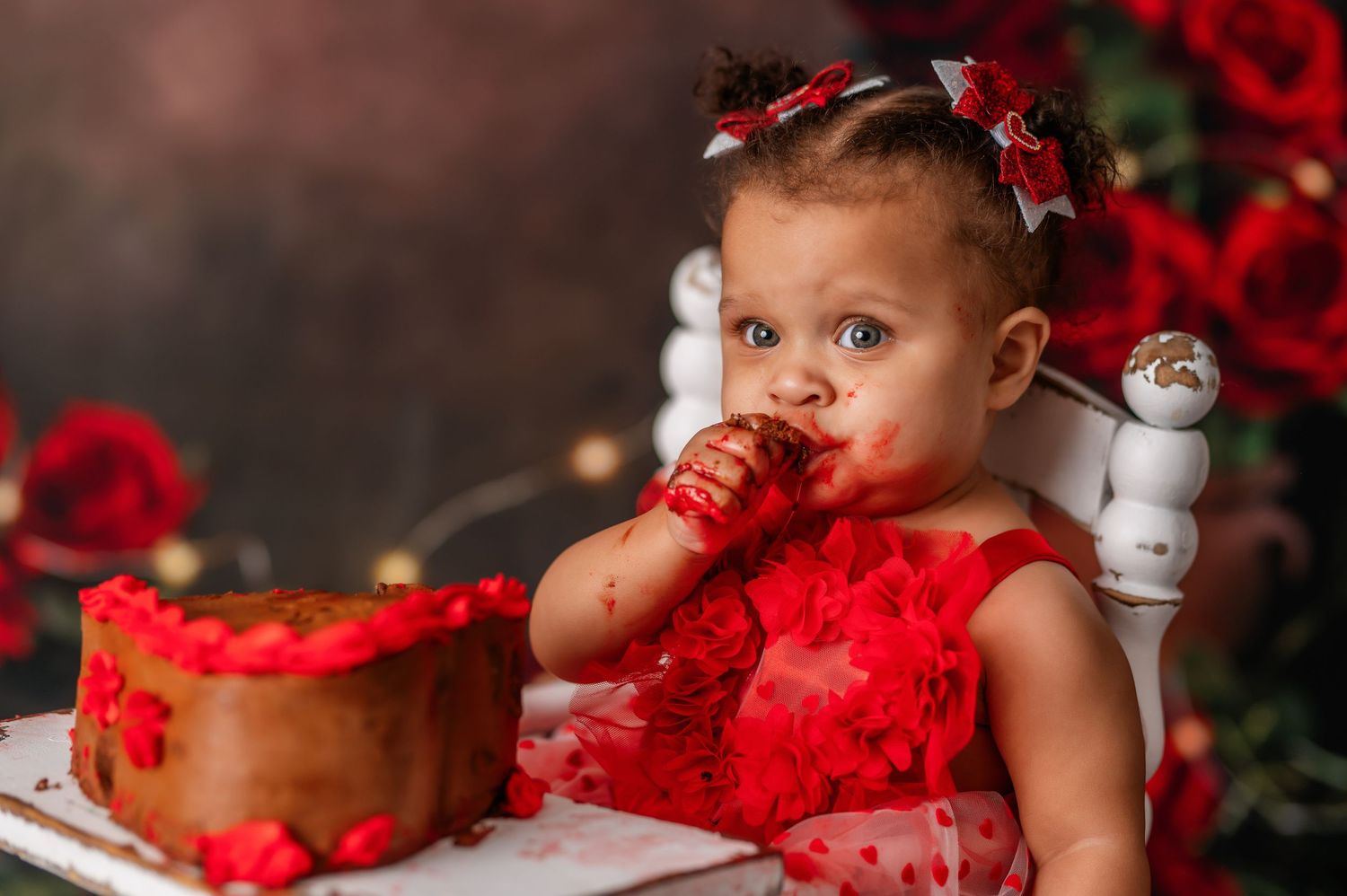 Baby enjoying cake during Valentine's themed photoshoot with red roses and tulle dress.