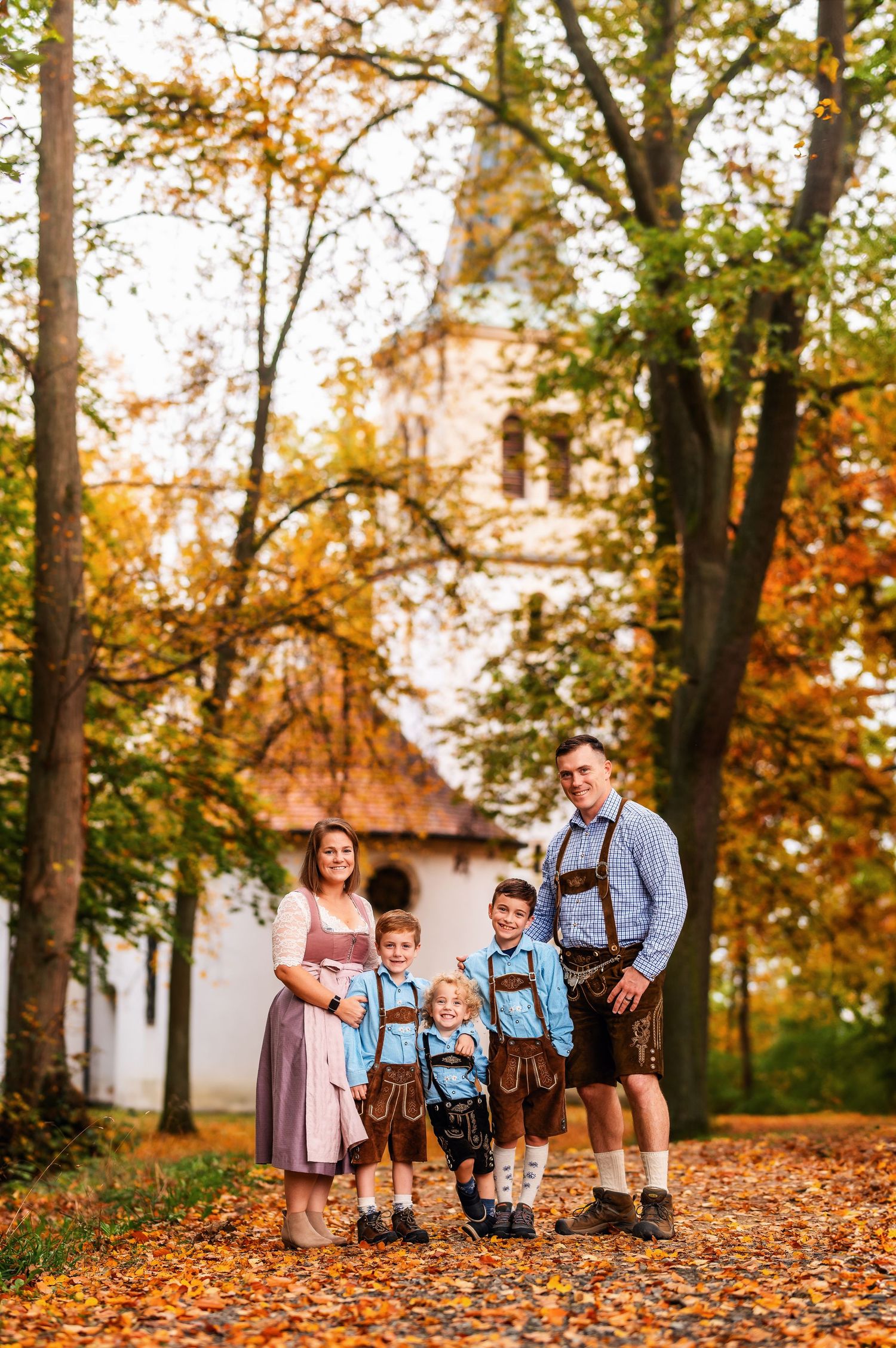 A family in traditional Bavarian dress poses among autumn trees near a white church.