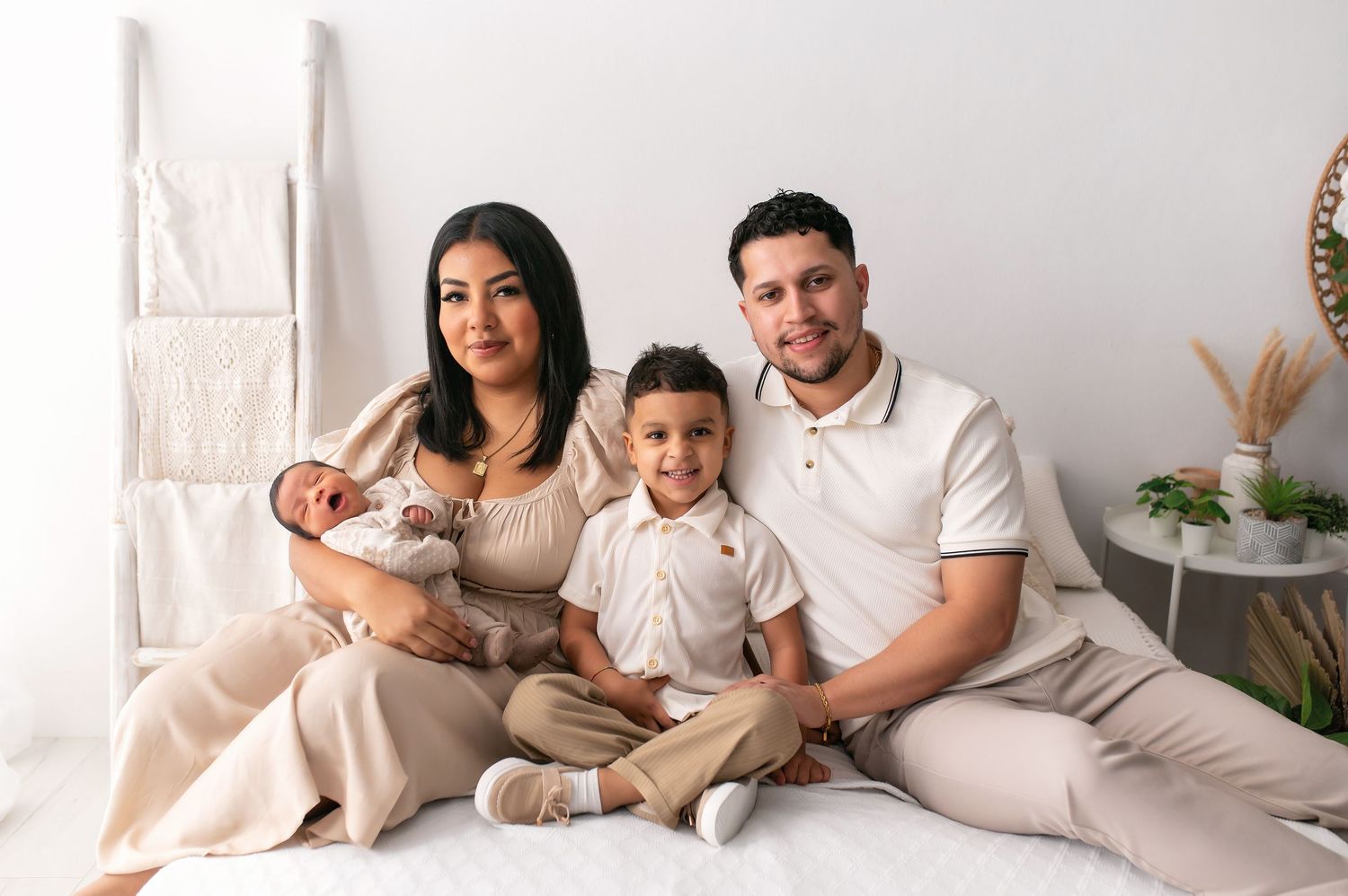Family of four poses for a portrait, mother holding newborn baby, young son seated between smiling parents in neutral tones.