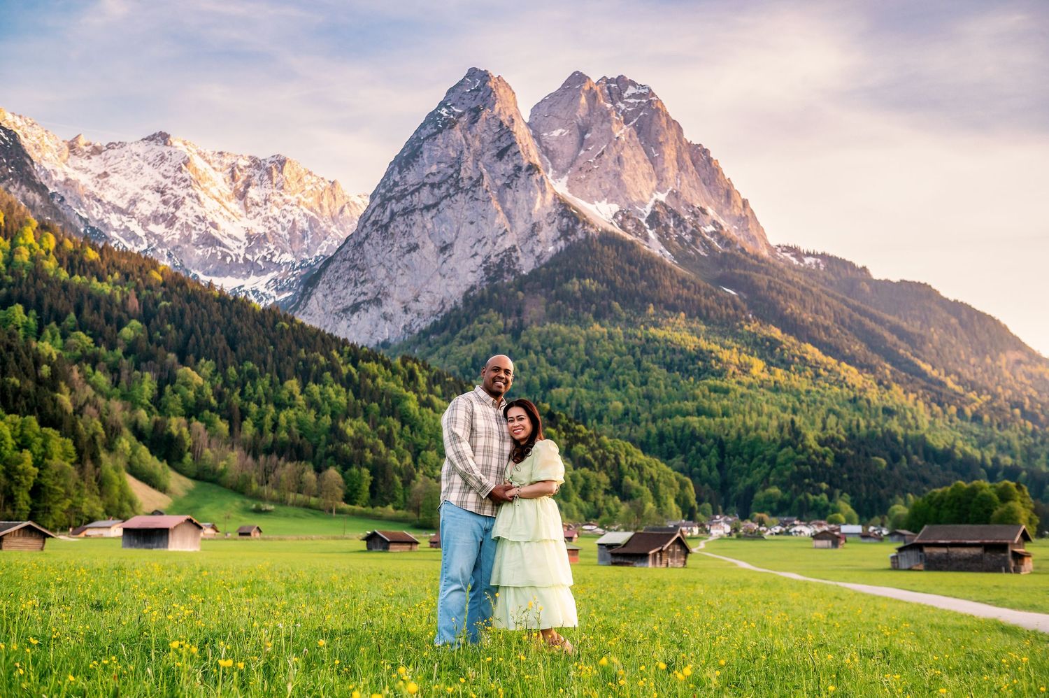 Couple embracing in a green alpine meadow with dramatic snow-capped mountain peaks and forested slopes at sunset.