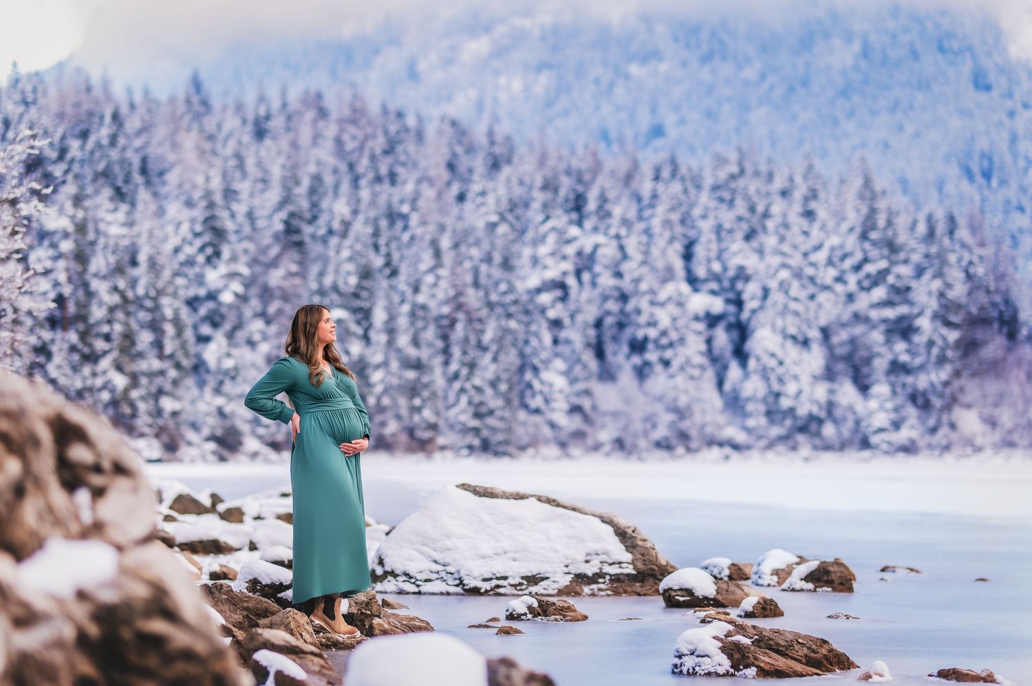 Person in teal dress stands on snowy lakeshore surrounded by snow-covered evergreen forest in winter landscape.