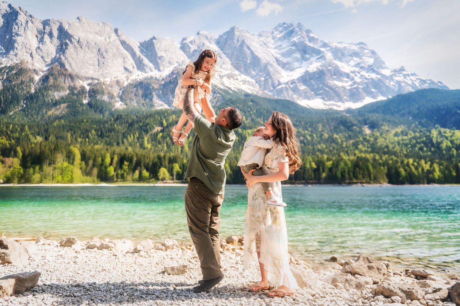 Family posing by a turquoise alpine lake with snow-capped mountains, father lifting child while mother holds baby.