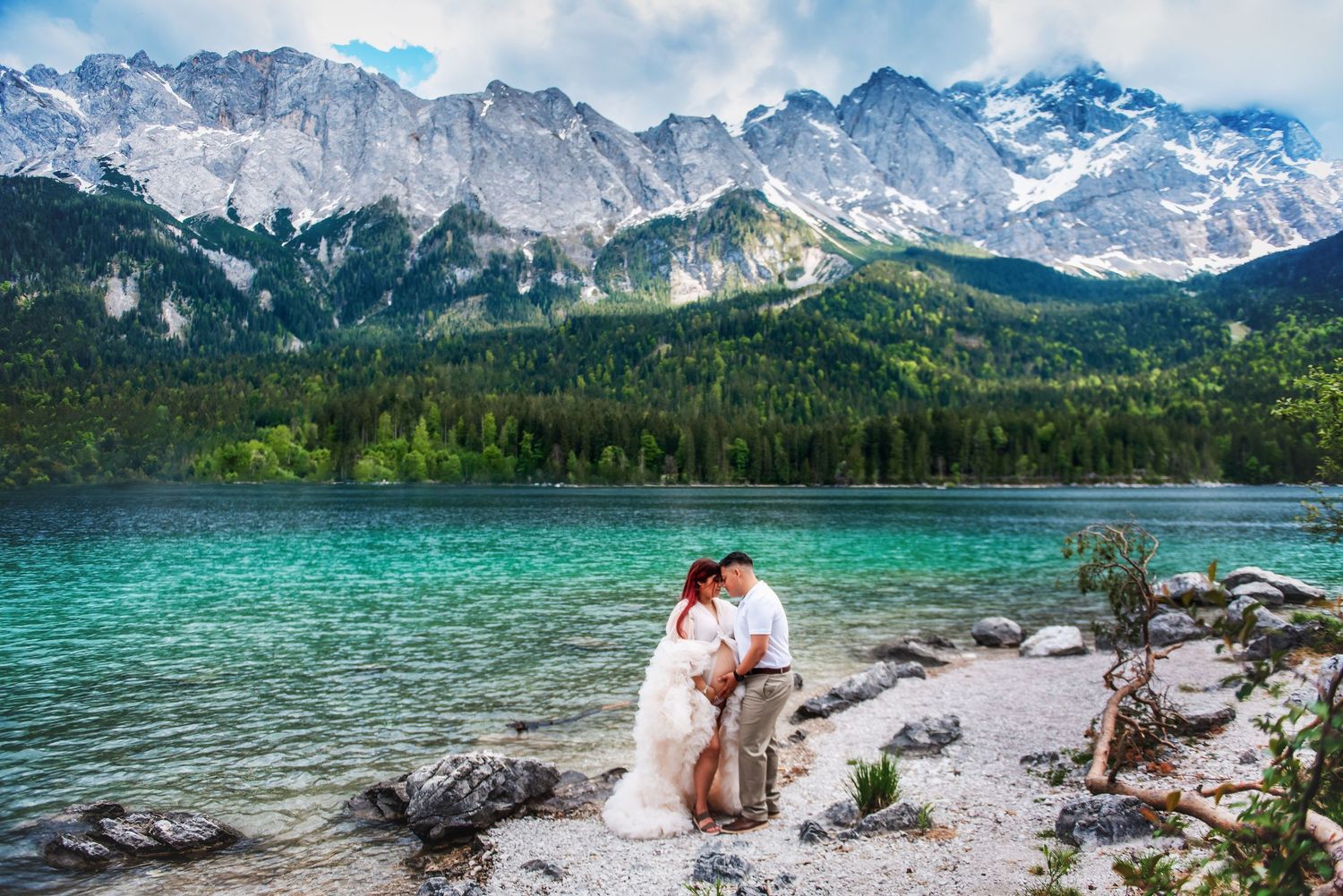 Couple embracing on rocky shore of turquoise alpine lake with snow-capped mountains and forest backdrop.