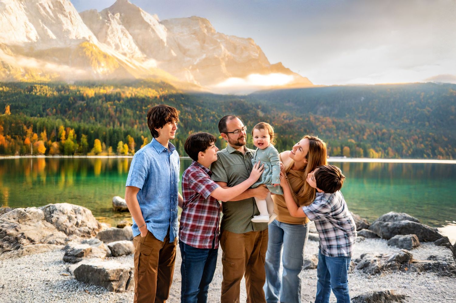 Family of six posing together by a turquoise mountain lake with autumn trees and rocky peaks in the background.
