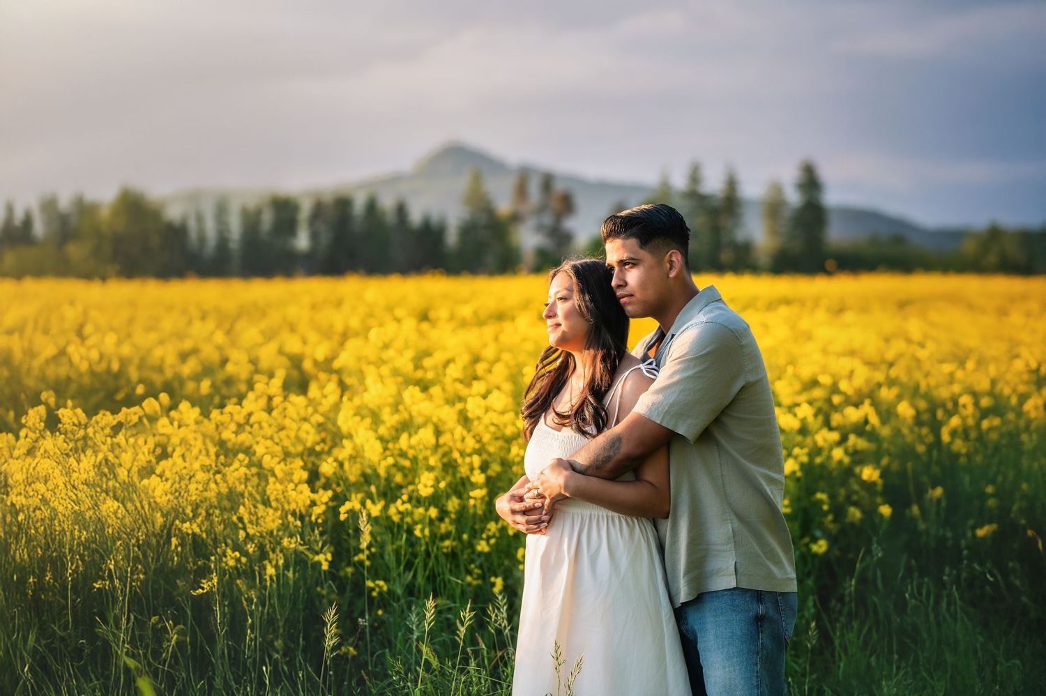 Romantic couple sharing intimate moments in a vibrant yellow canola field with mountains in the background.