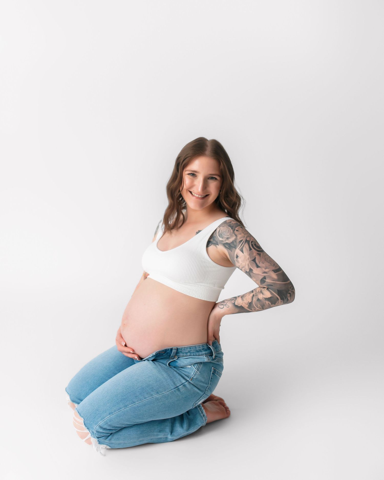 Pregnant woman with tattoos kneels in casual wear during studio photoshoot.