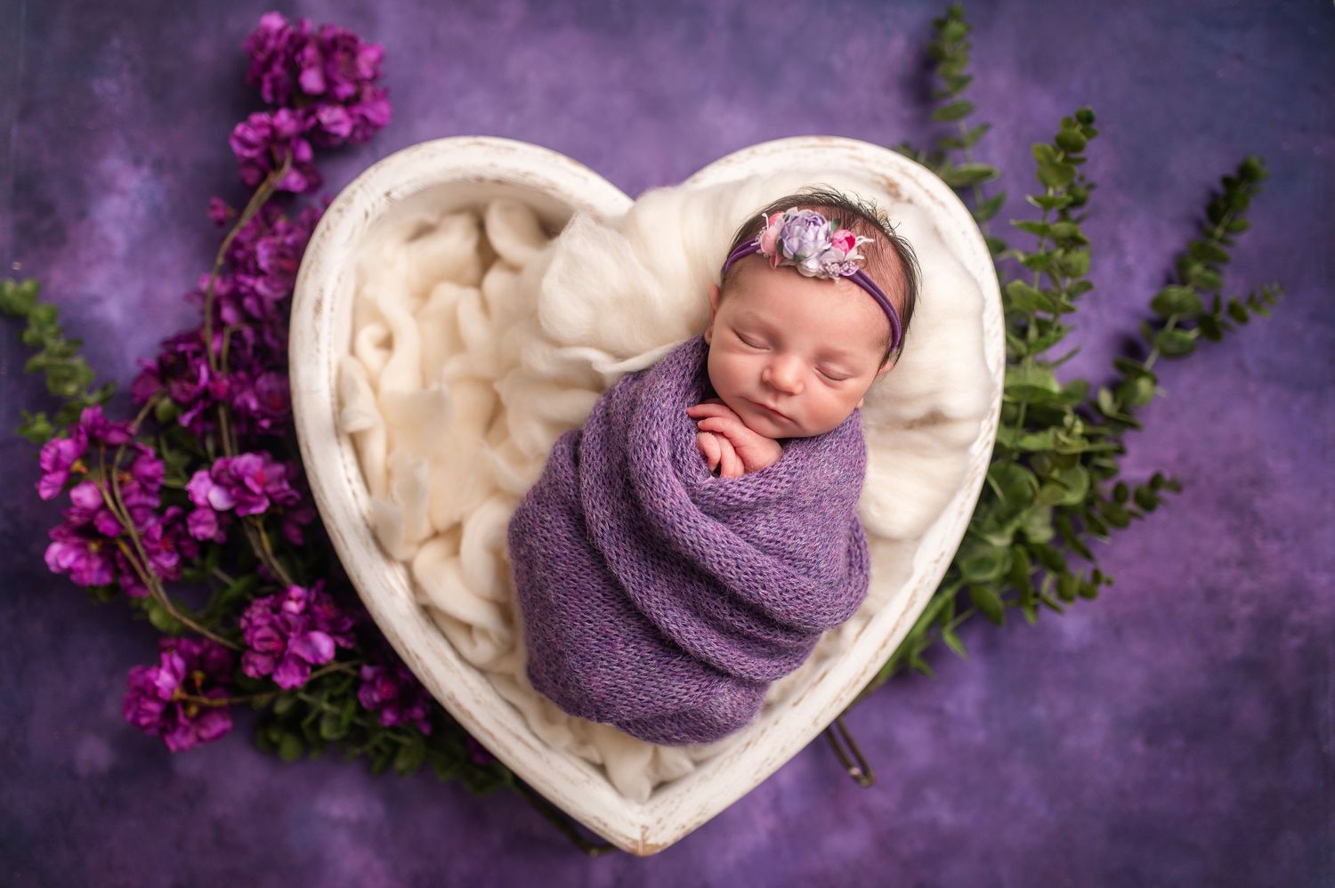 Sleeping newborn wrapped in purple swaddle in heart-shaped bed with floral accents.