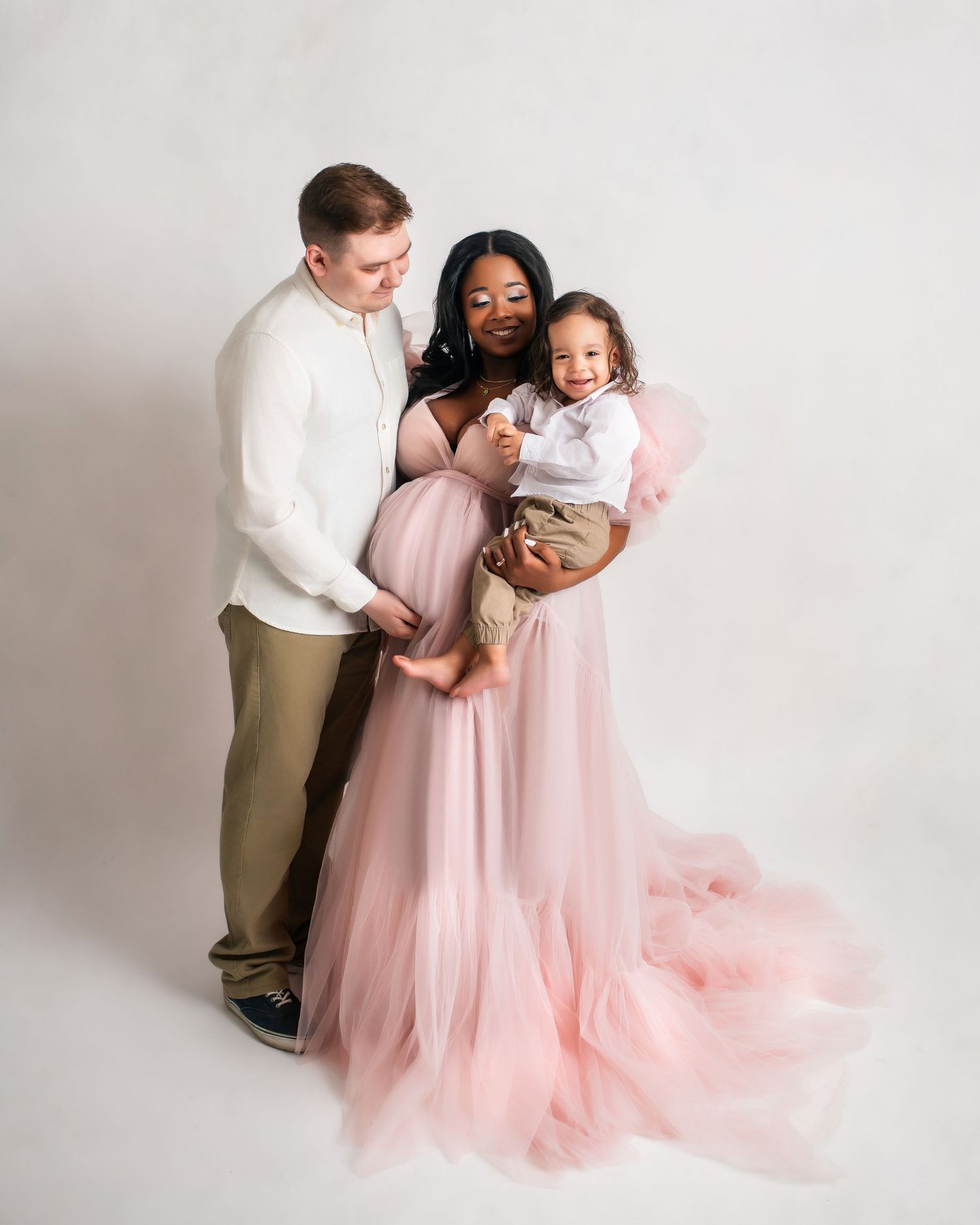 Studio maternity portrait features a family in soft pink and white attire against a light backdrop.