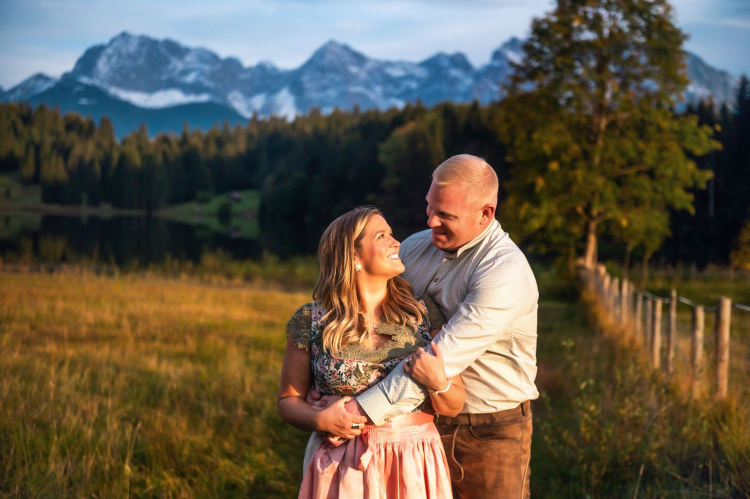A couple embraces in traditional dirndl and lederhosen against a scenic mountain backdrop at sunset in rural Bavaria.