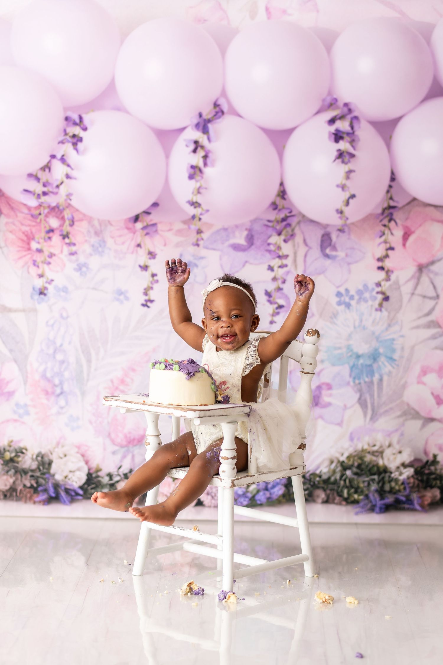 Baby enjoying cake smash photo session on white chair against pink balloon and floral backdrop.