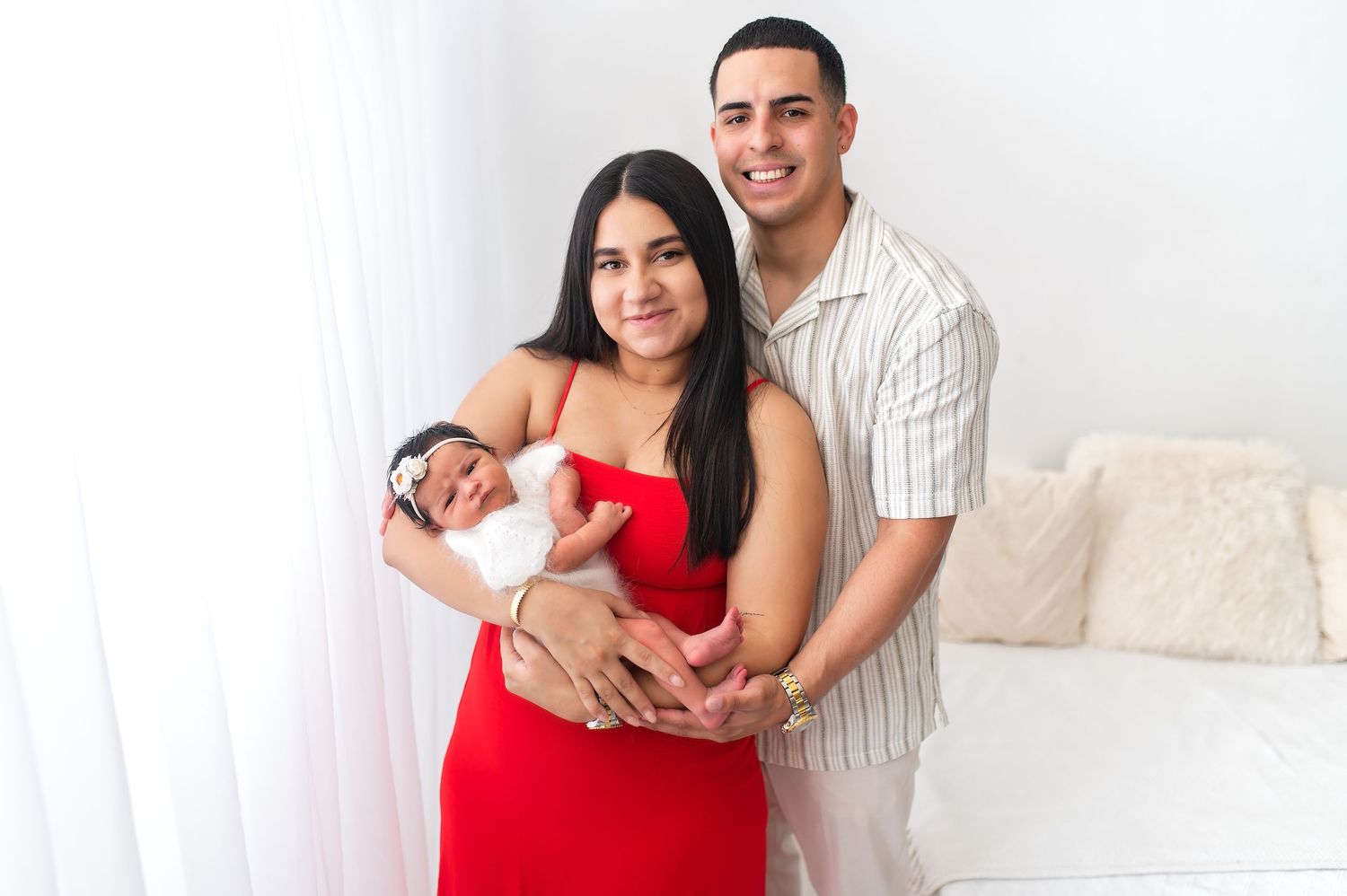 New parents in red dress and striped shirt hold their newborn baby in bright home setting.