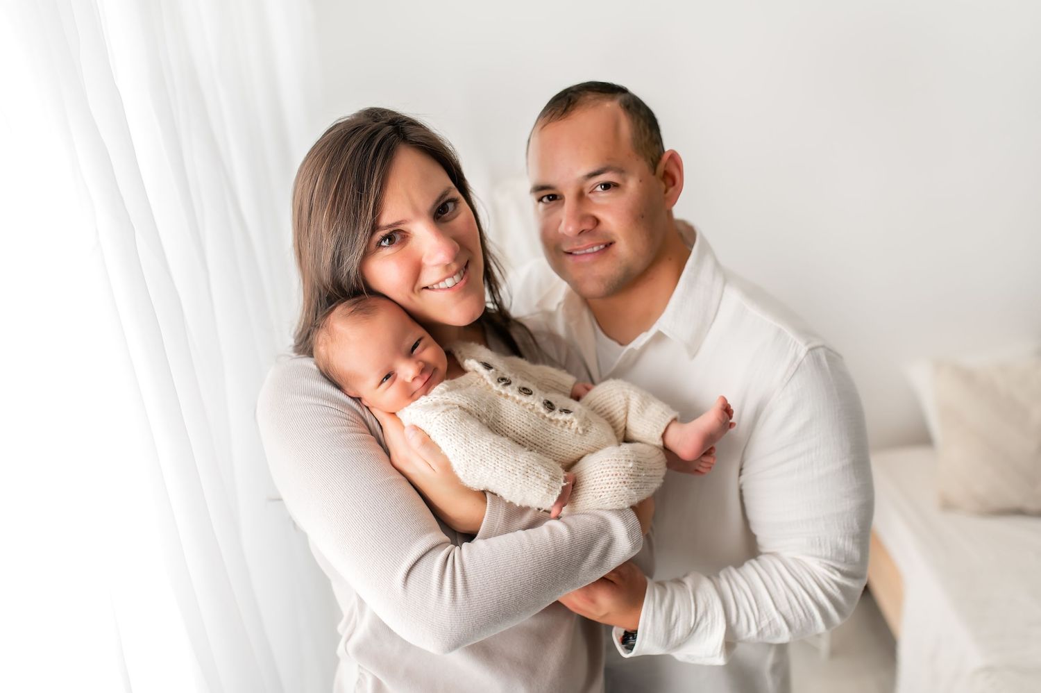 Parents holding newborn baby in neutral toned indoor setting.