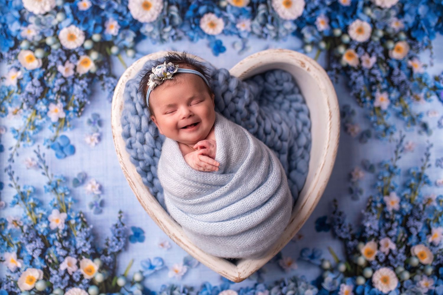 Newborn wrapped in gray swaddle in heart-shaped prop with blue floral background.