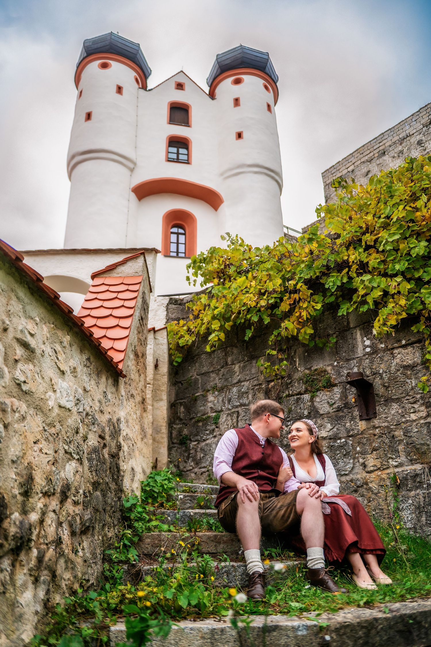 A romantic scene in front of a historic castle with white towers and red tiled roofs surrounded by stone walls and greenery.