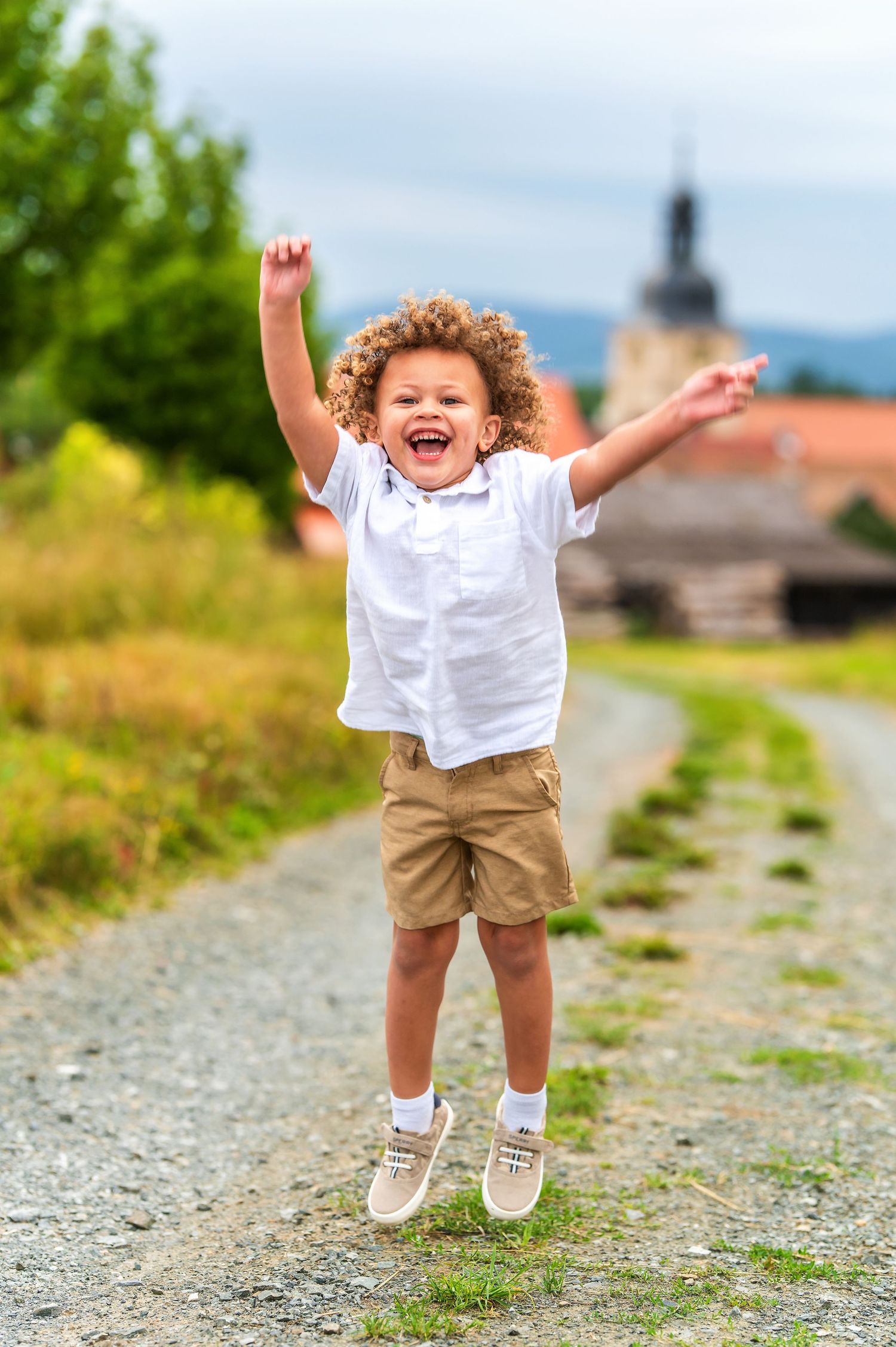 Child in white shirt and shorts jumps joyfully on rural path with village in background.