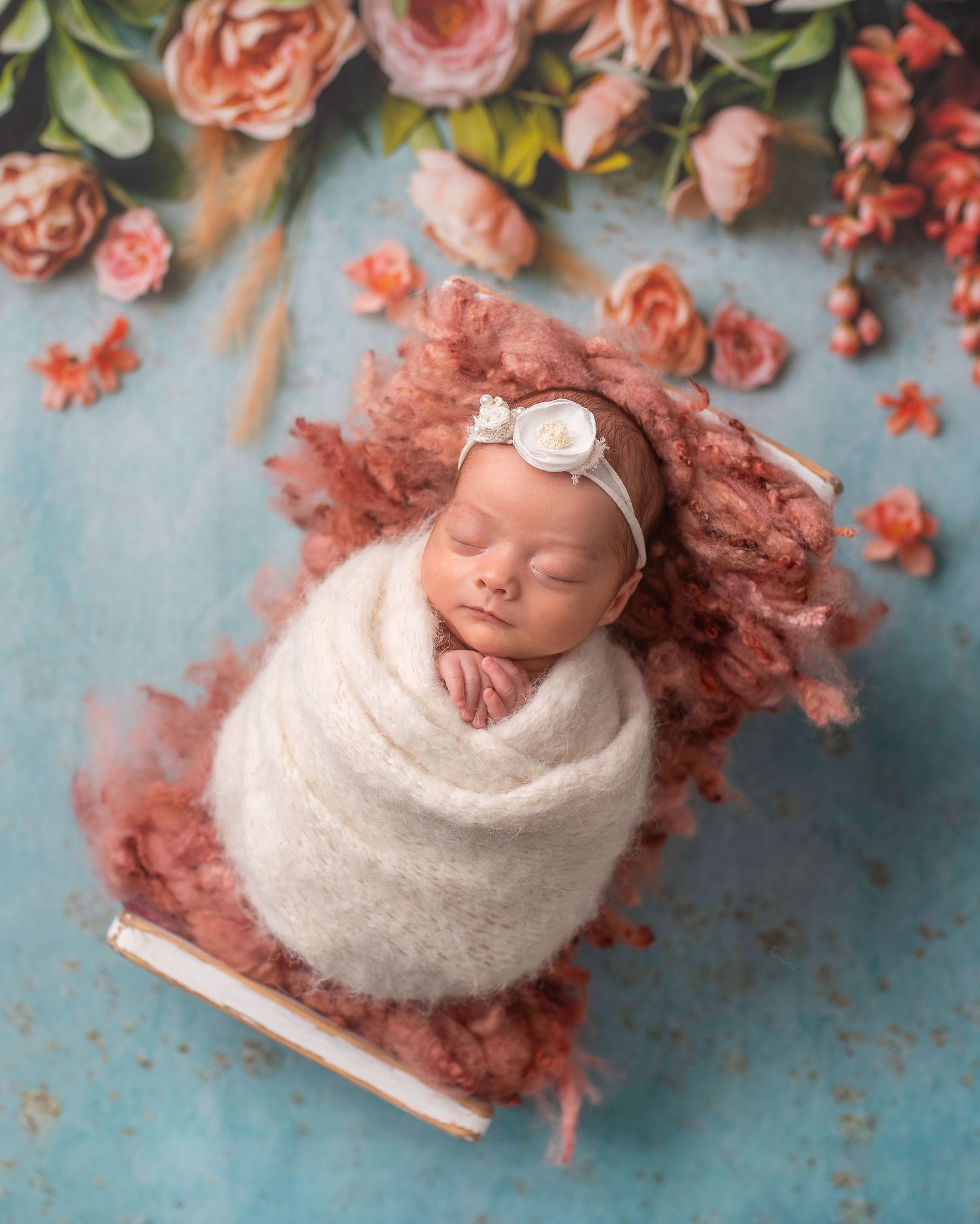 Newborn baby swaddled in cream wrap with floral headband, nestled in pink fluff surrounded by pink roses on teal background.